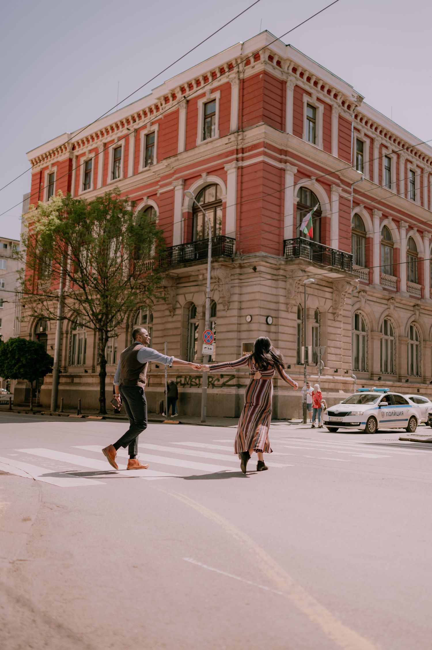 Couple holding hands while crossing a zebra crossing in front of a grand red historic building on a sunny day.