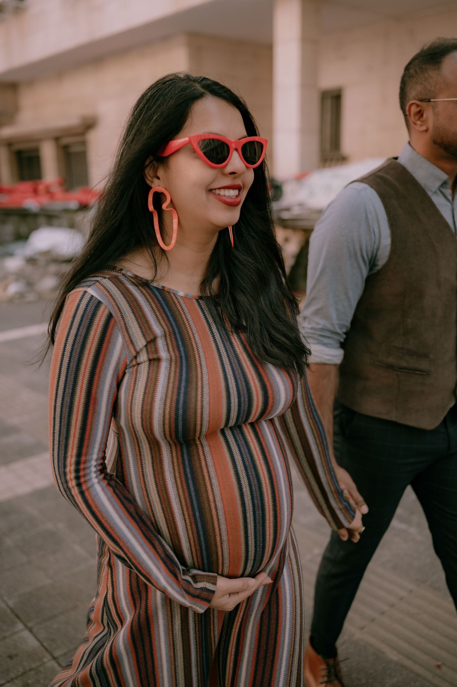 Smiling pregnant woman in striped dress and red cat-eye sunglasses with hoop earrings walking outdoors.