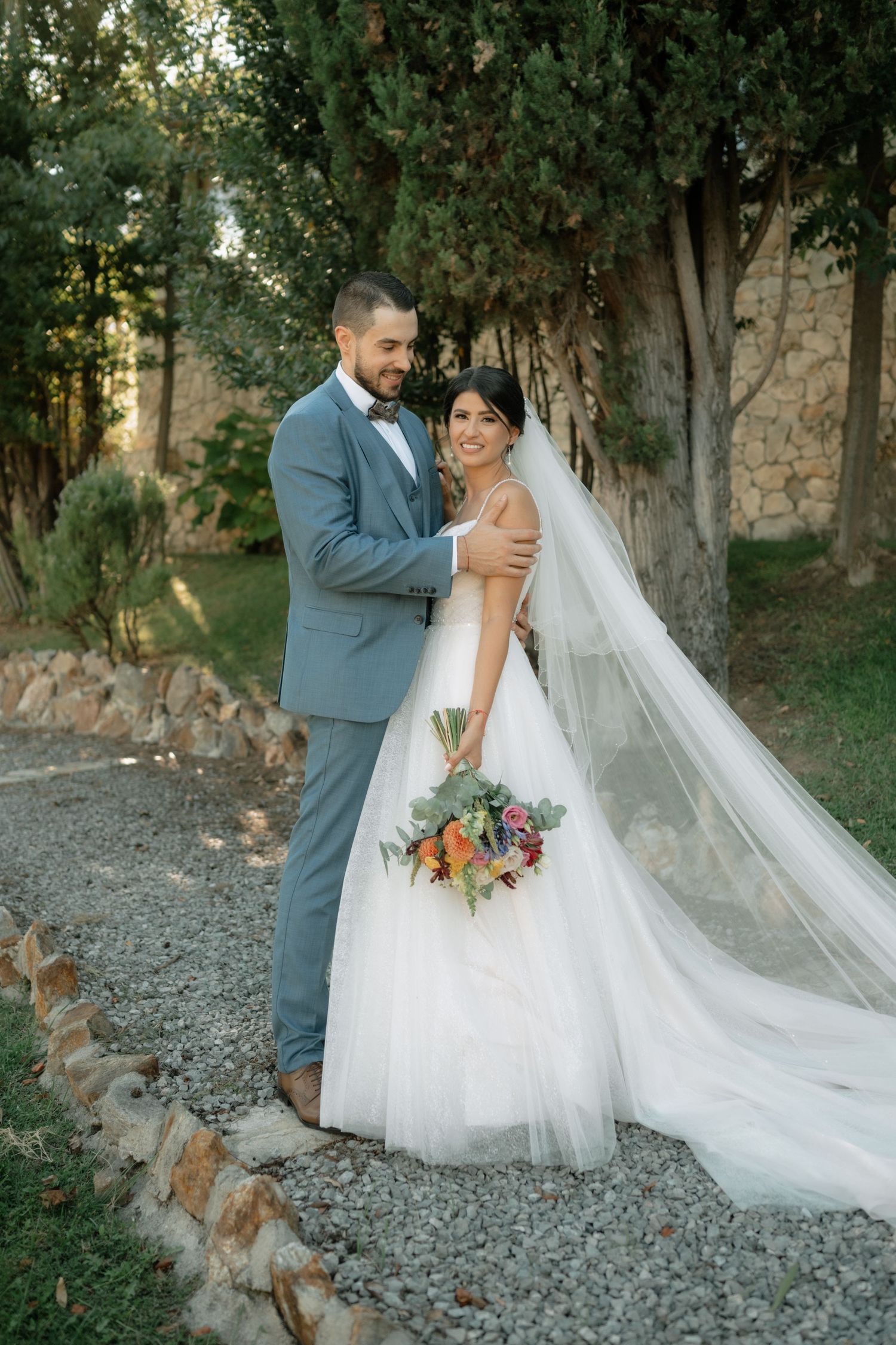 A couple in wedding attire poses together outdoors with a flowing white veil and colorful bouquet against a wooded backdrop.