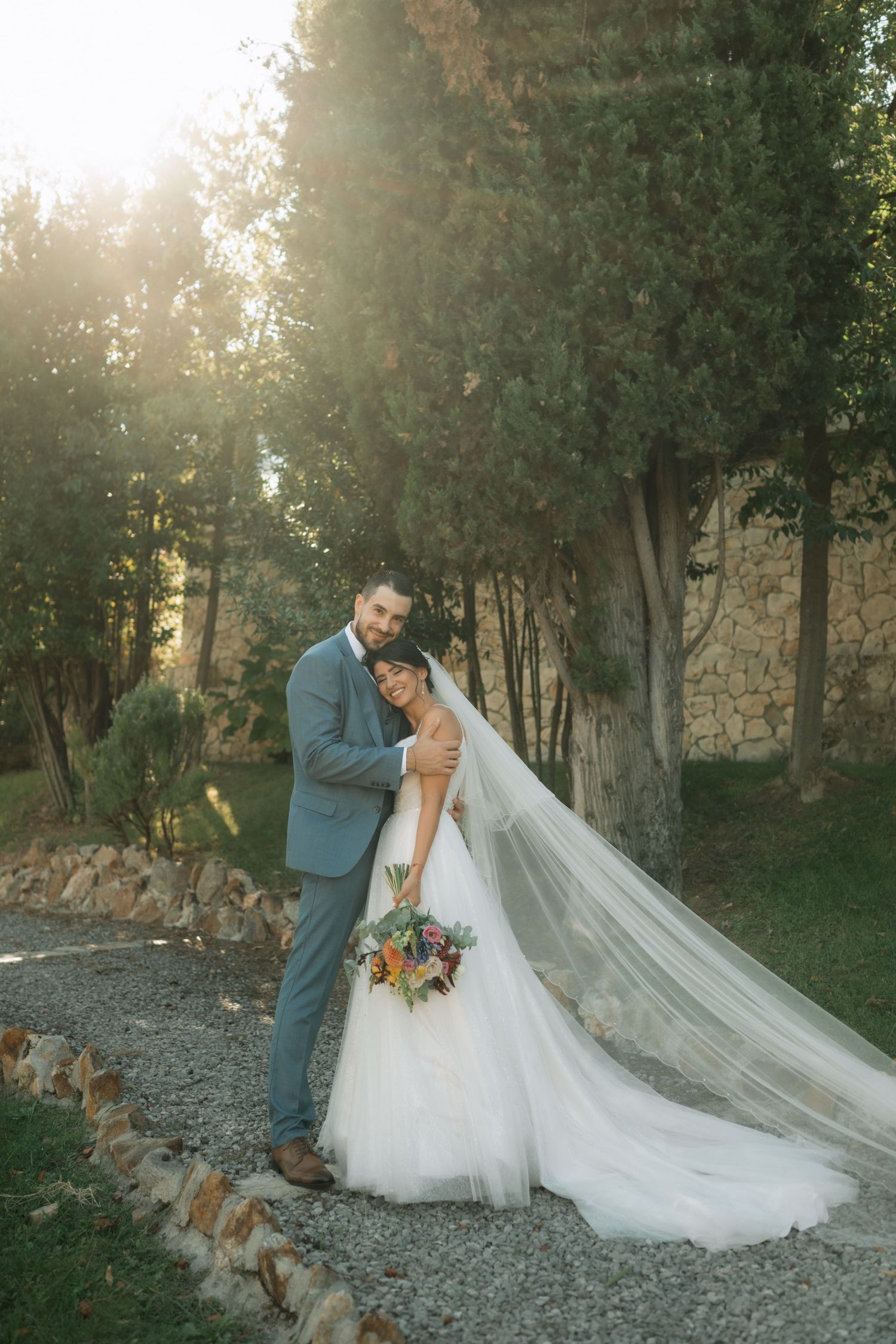 A couple in wedding attire poses together on a rocky path surrounded by trees during golden hour.