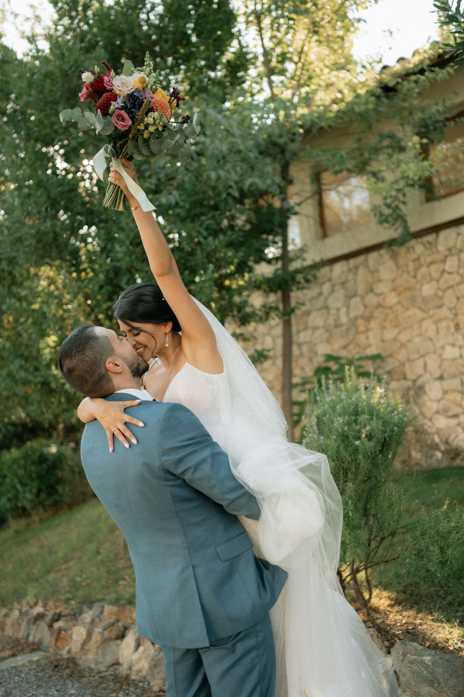 A romantic wedding couple embraces and dances together outdoors against a rustic stone wall at sunset.