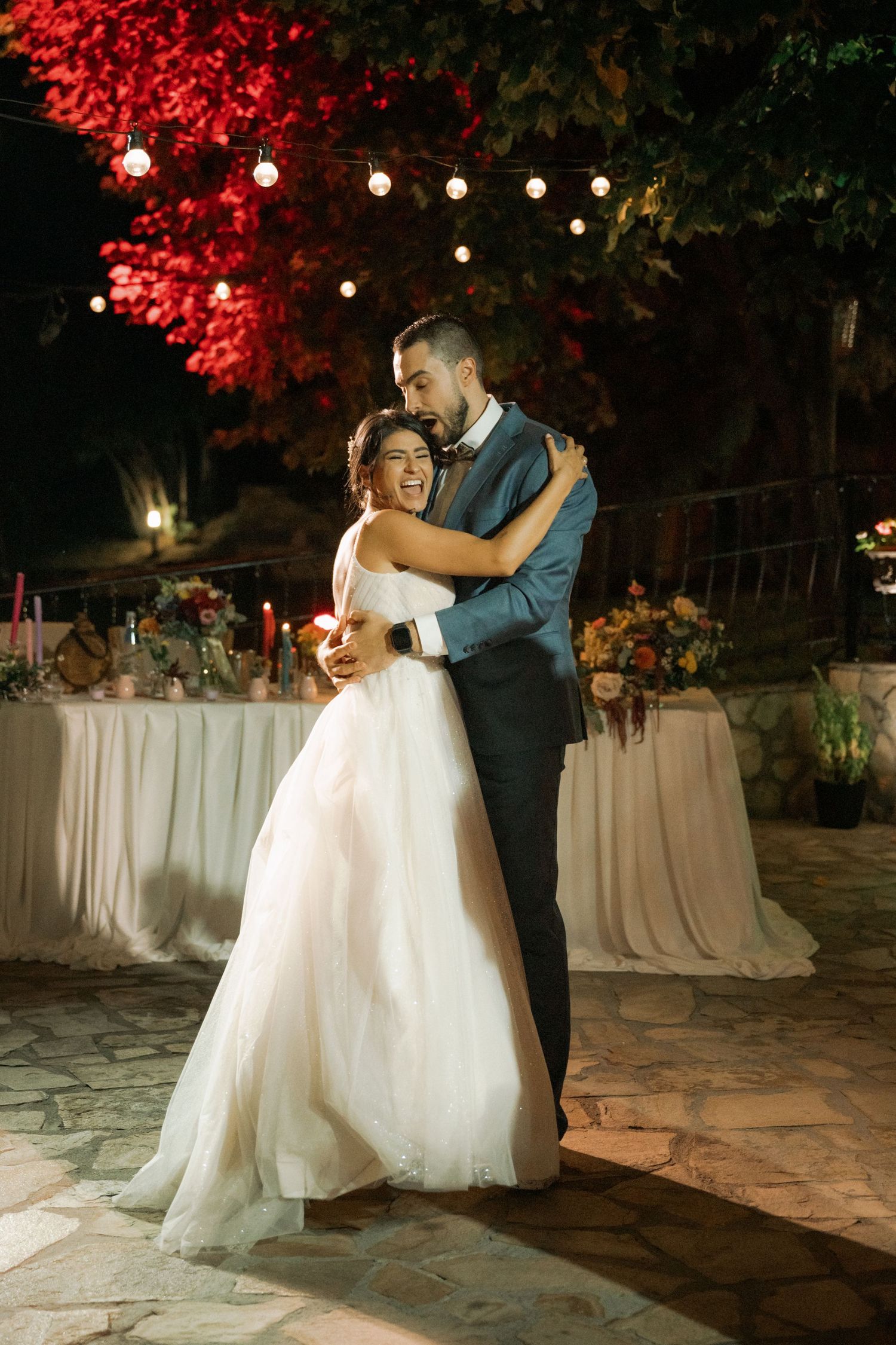 A couple shares their first dance at an outdoor evening wedding reception under red autumn foliage and string lights.