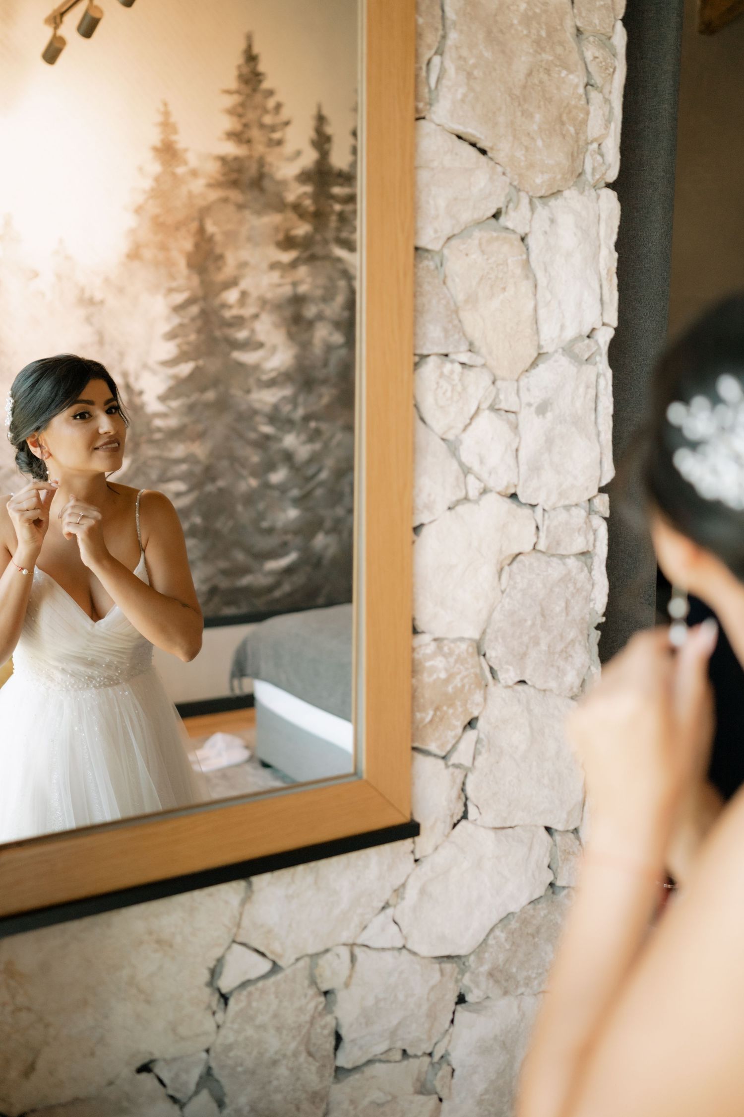 A reflection in a wooden-framed mirror shows a white dress against a stone wall with pine trees visible in the background.