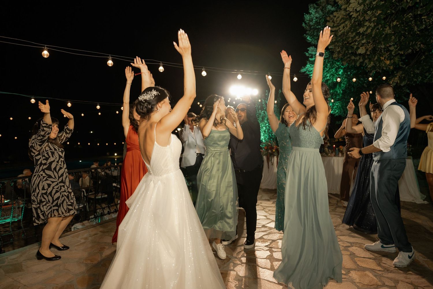 Wedding guests dancing and celebrating on an outdoor dance floor at night under string lights during a reception party.