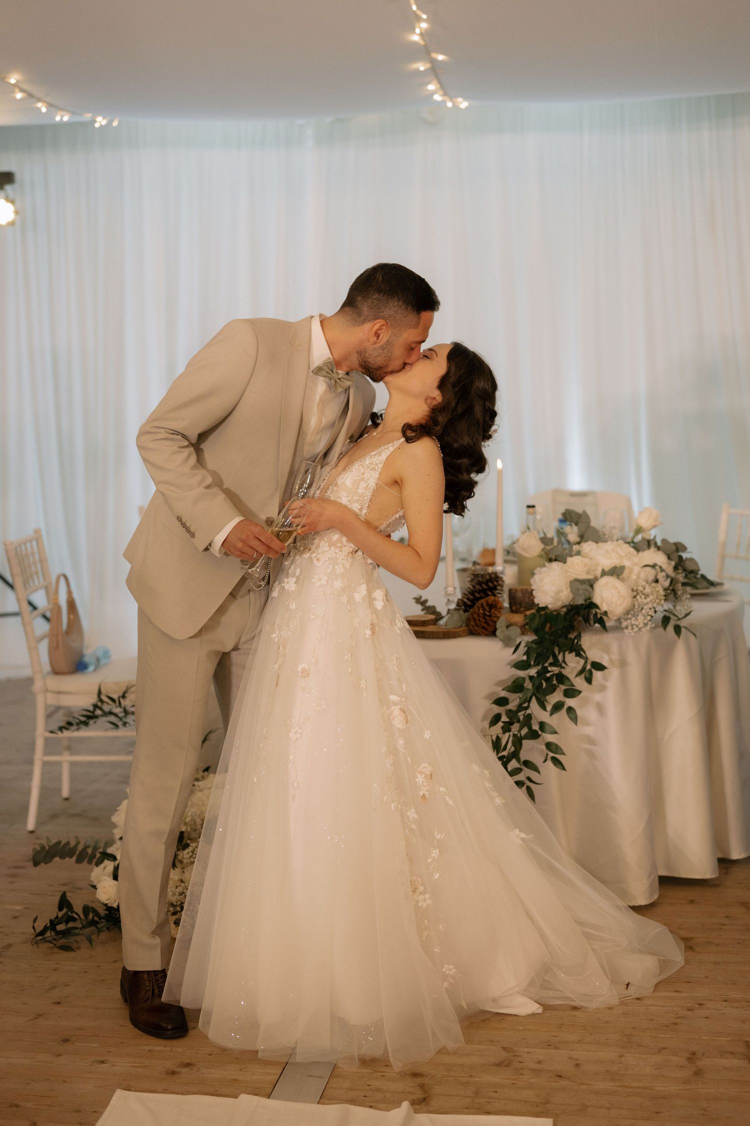 A bride and groom share a romantic kiss at their wedding reception, with white drapes and fairy lights in the background.