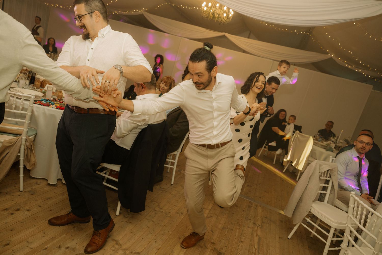 Men dancing energetically at a wedding reception in a decorated venue with white chairs and draped ceiling lights.