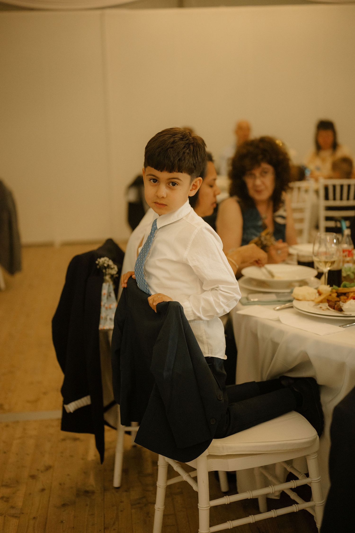 Young boy in white shirt and blue tie holds suit jacket at a formal wedding reception dinner table.