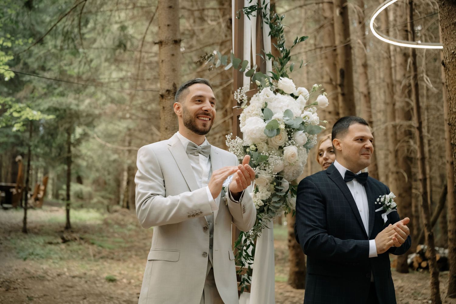 Groom in beige suit clapping at outdoor forest wedding ceremony with floral arch.