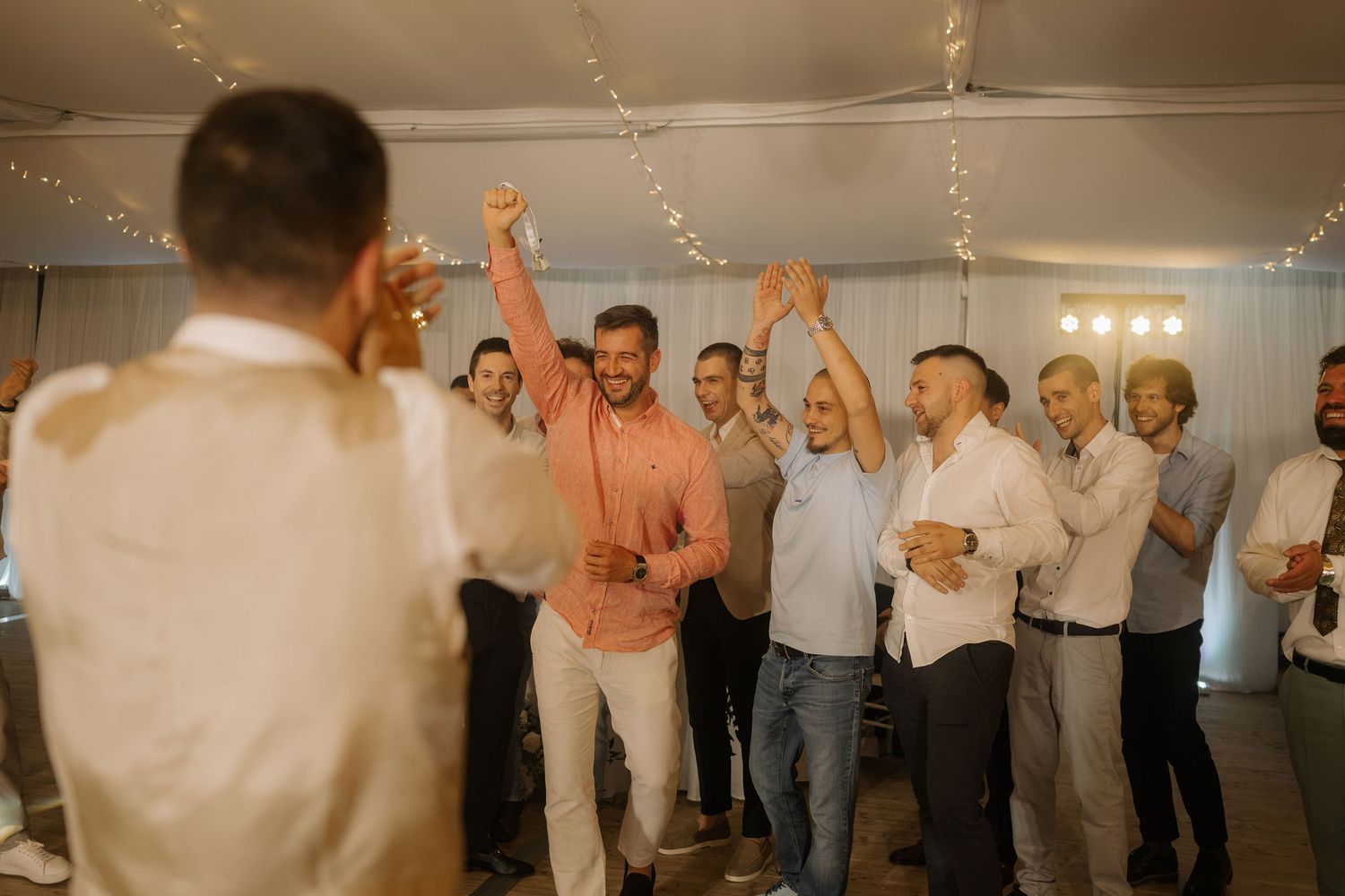Men celebrating at a wedding reception, raising glasses and dancing under string lights in a white tent.