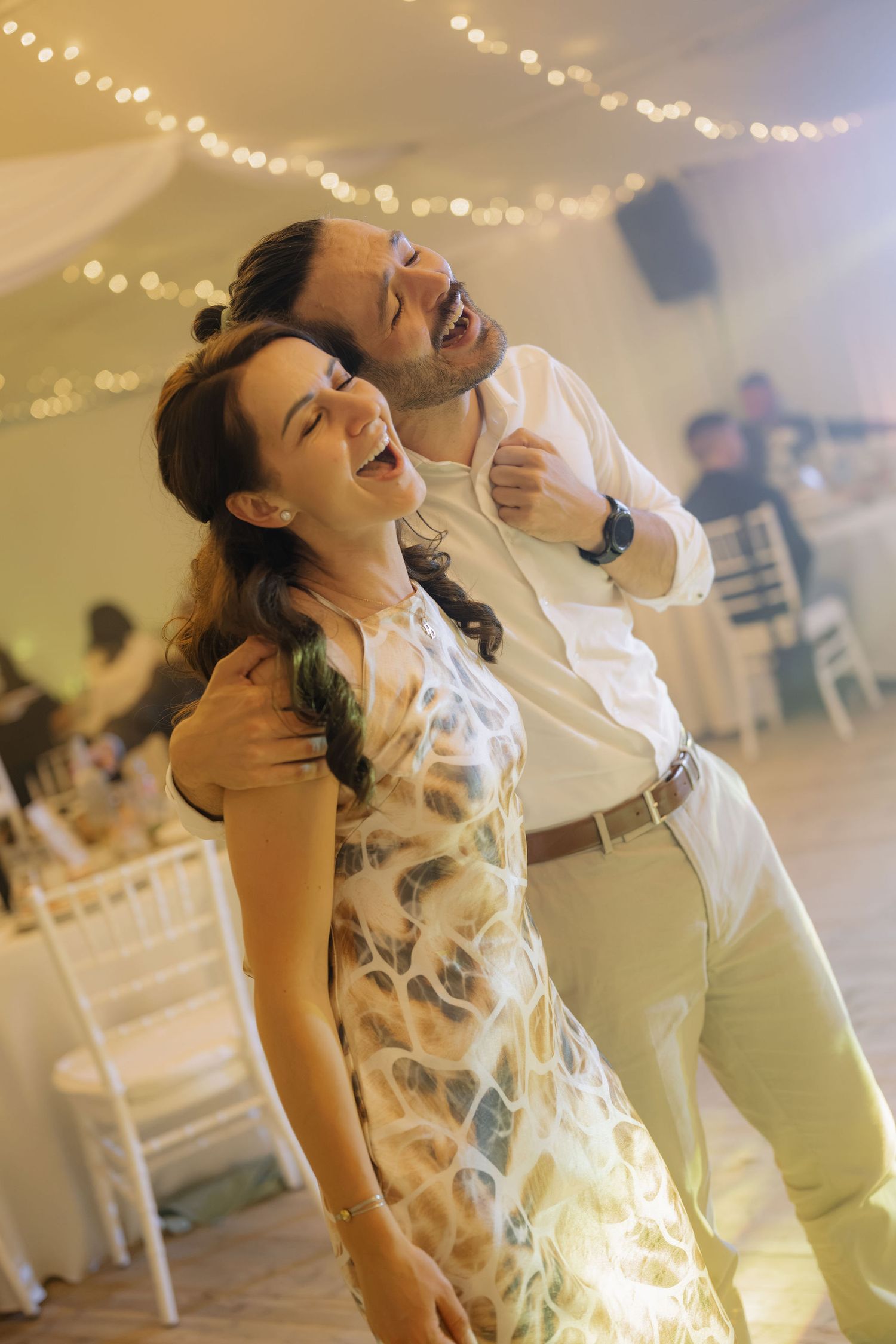 A joyful couple laughing and dancing together at an elegant wedding reception with warm string lights.