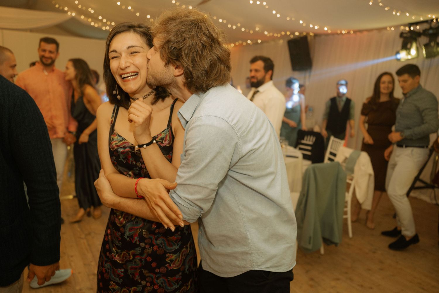 Couple dancing and laughing together at a wedding reception with string lights and guests in background.