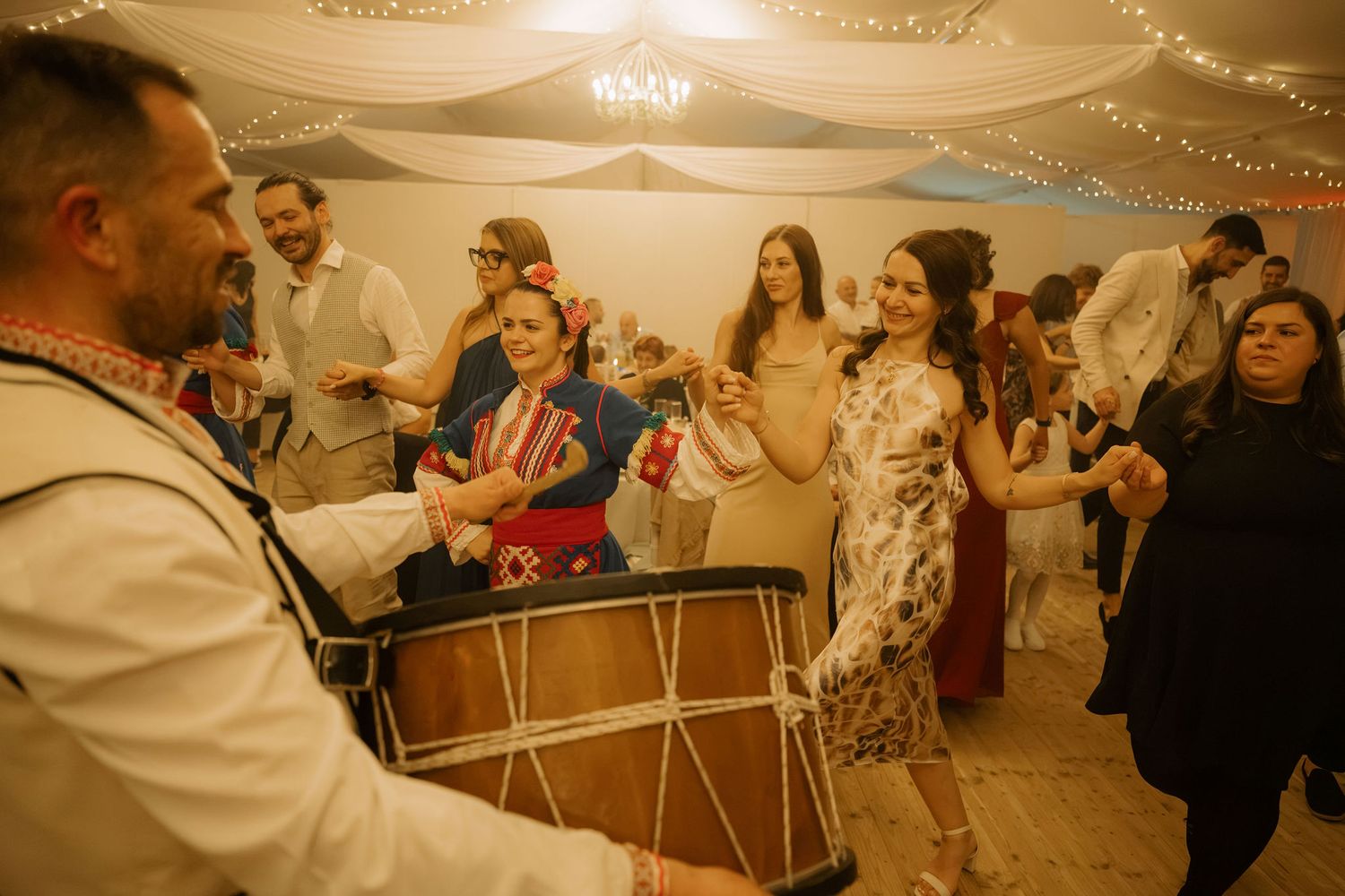 Guests dancing joyfully at a wedding reception while a musician plays a traditional drum indoors.
