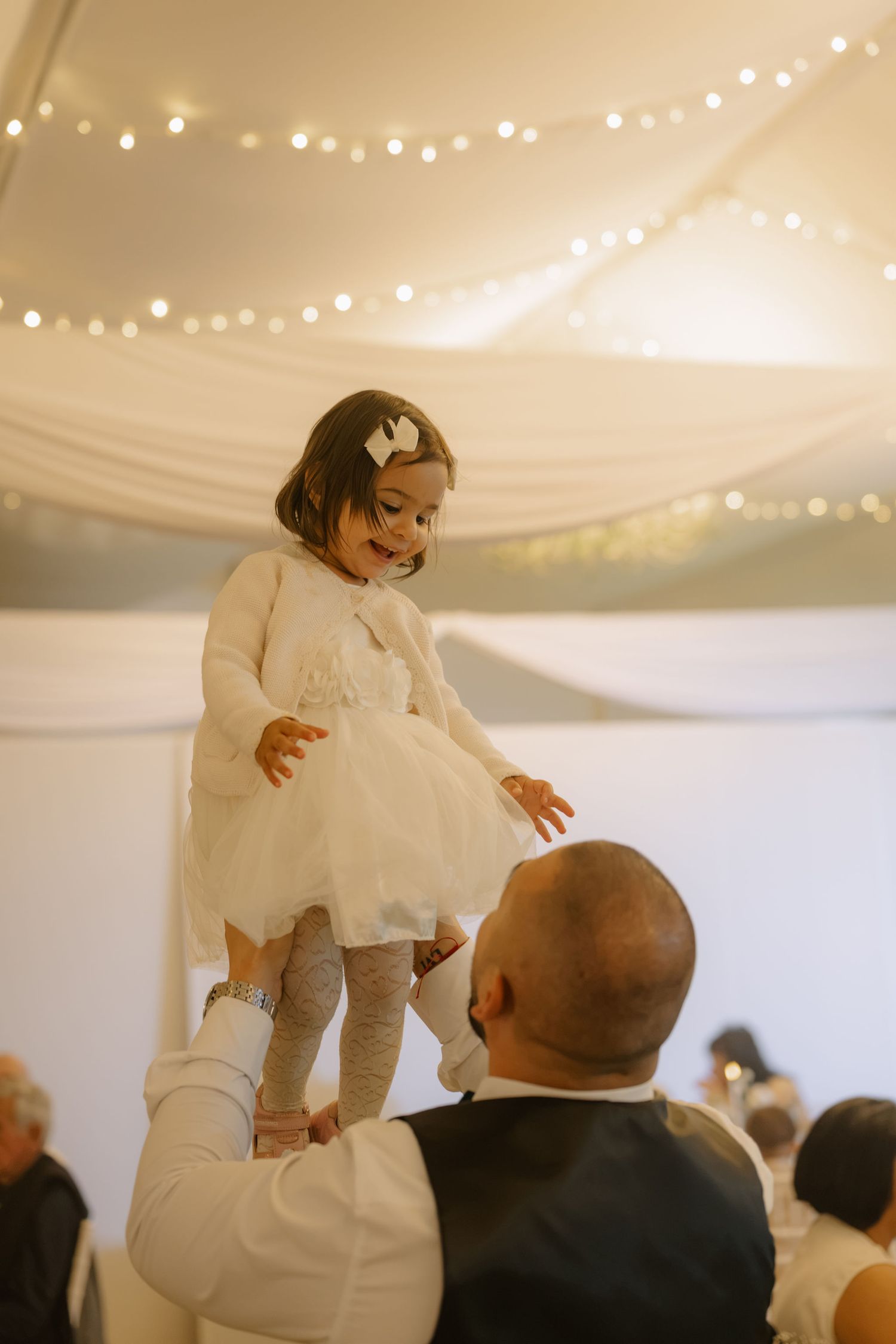 A man lifts a young girl in a white dress at a wedding reception with fairy lights on the ceiling.