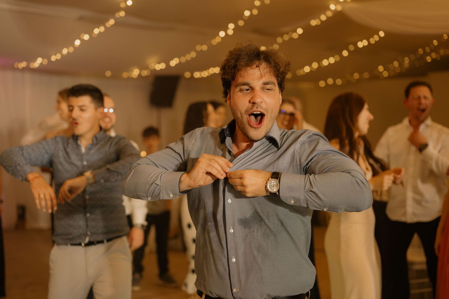 Man dancing energetically at a wedding reception, making a funny face, with string lights and guests in the background.