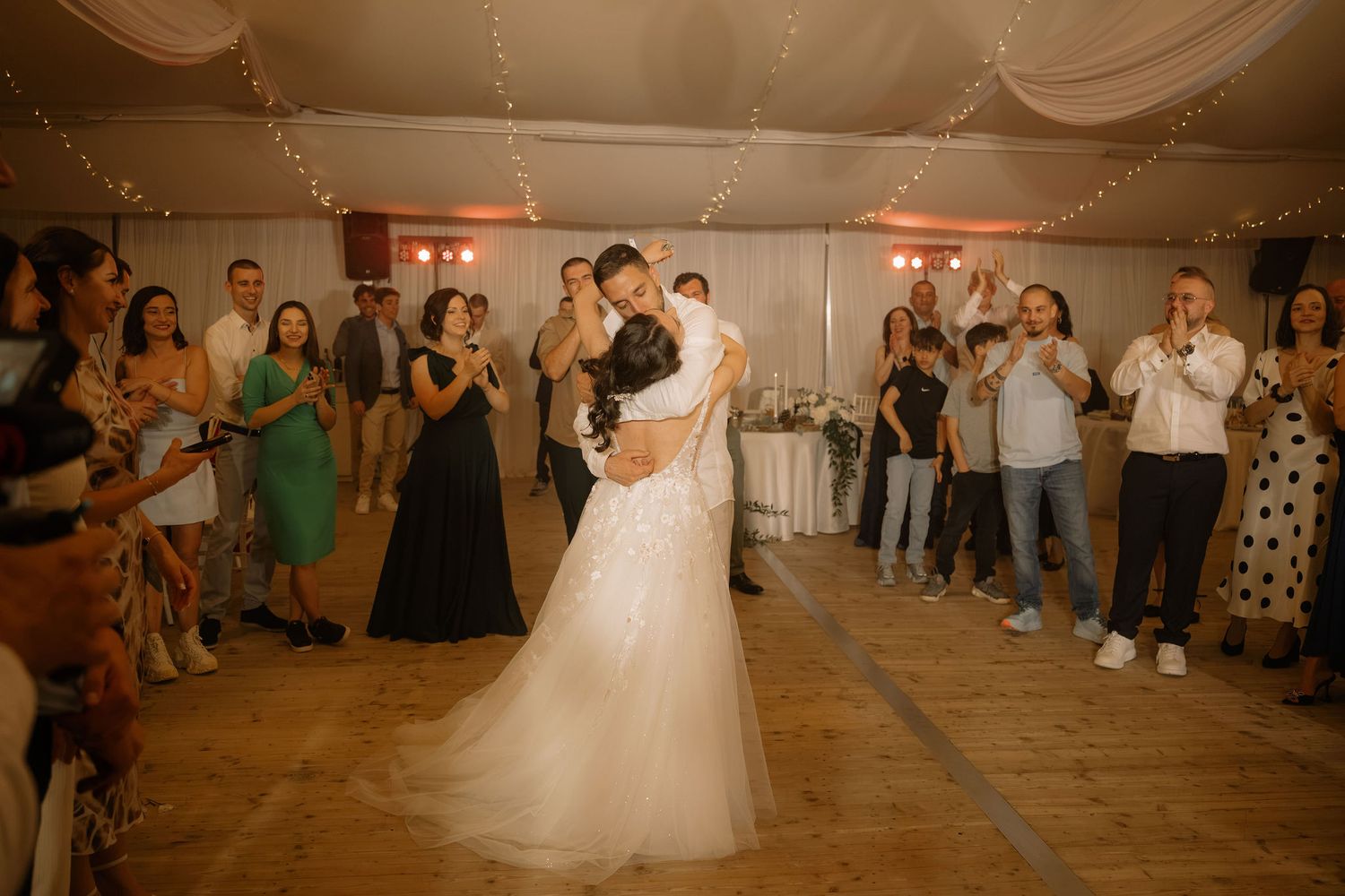 Bride and groom share first dance at wedding reception surrounded by applauding guests under fairy lights.