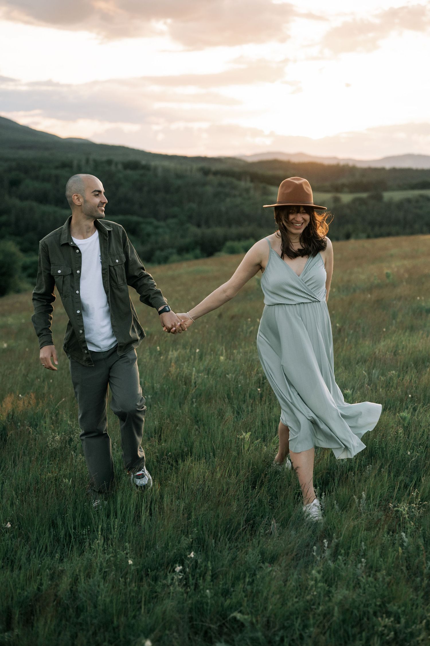 Couple holding hands walking through green meadow at sunset, woman in light dress and hat, man in olive jacket.