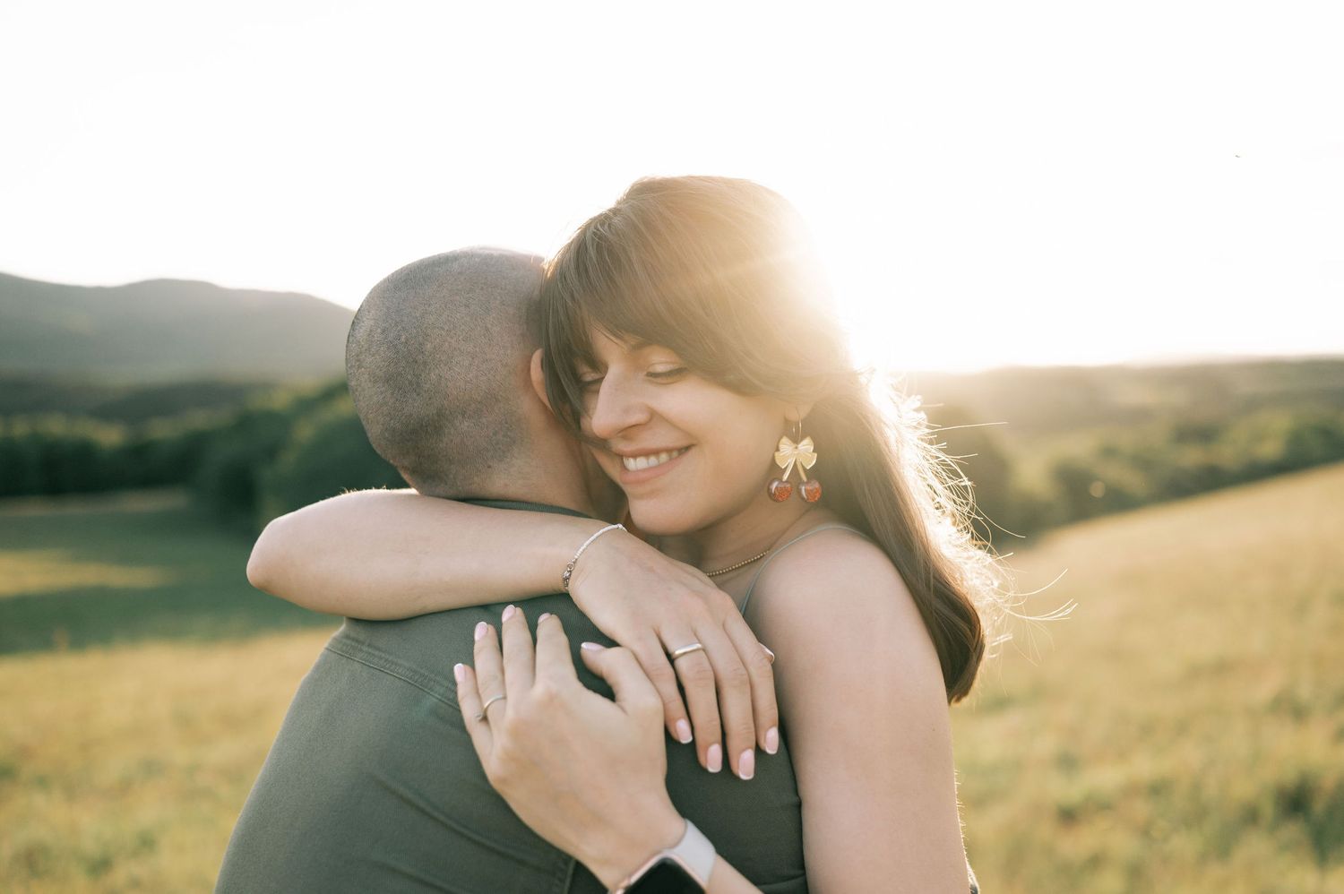 Couple embracing in golden sunset light in a green field, woman smiling with engagement ring visible.