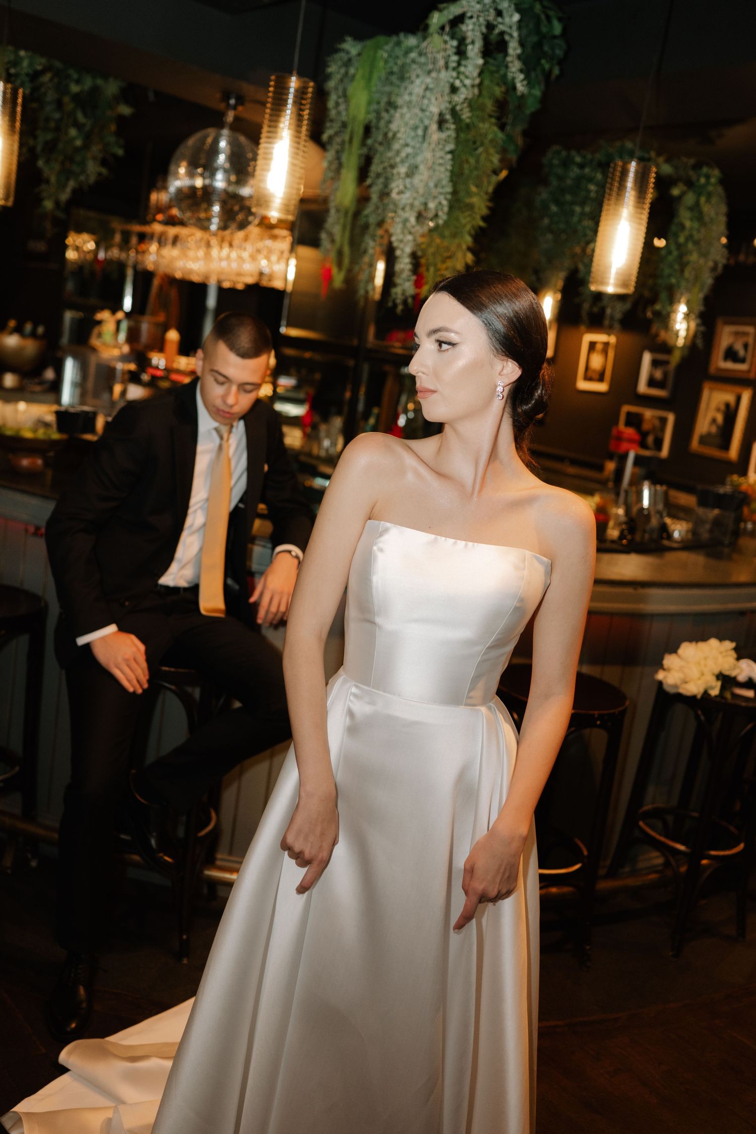 A bride in a white strapless wedding gown stands in an elegantly lit reception venue at night.