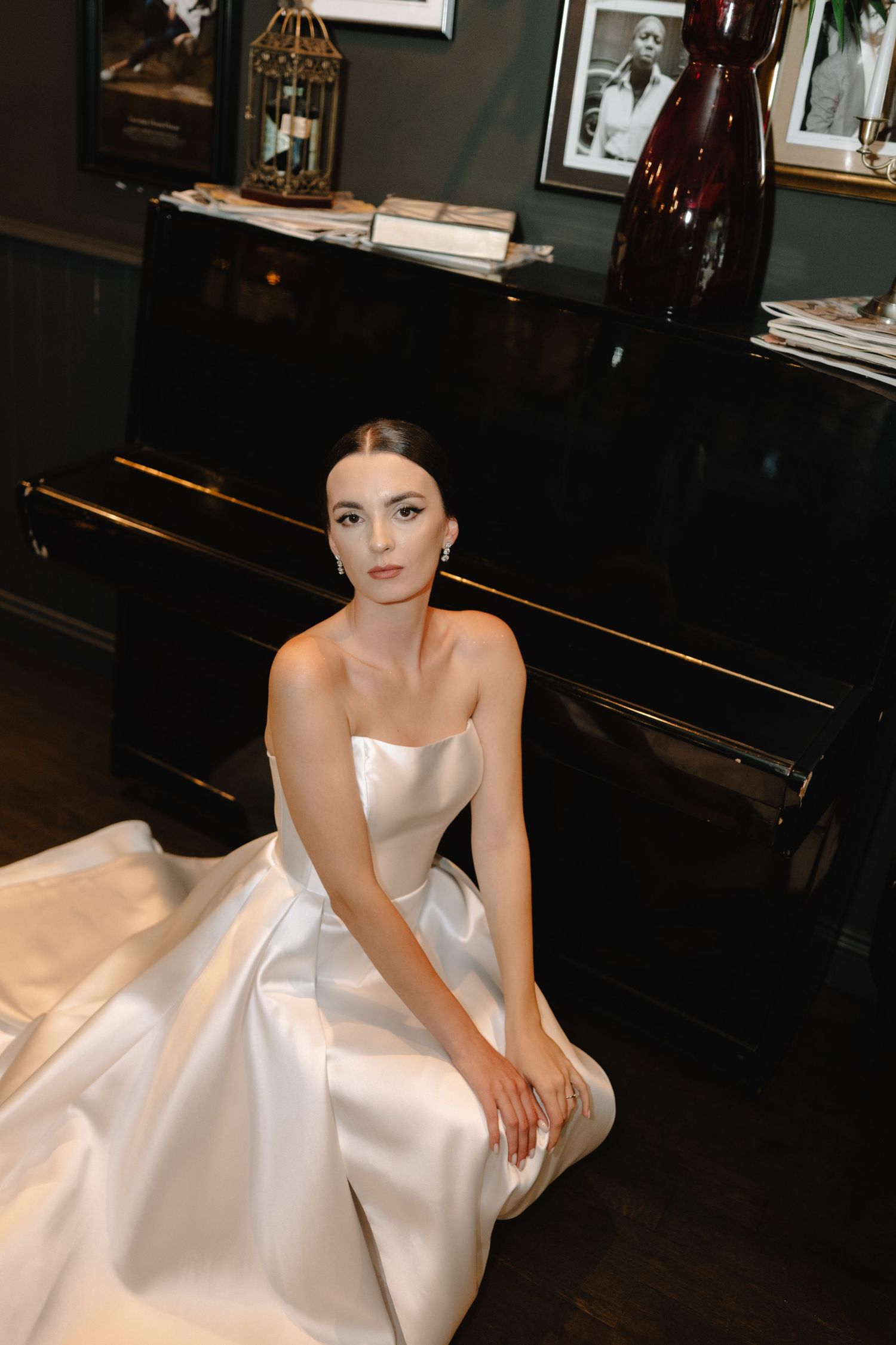 Elegant strapless white satin wedding gown with full skirt photographed in dark room with piano.