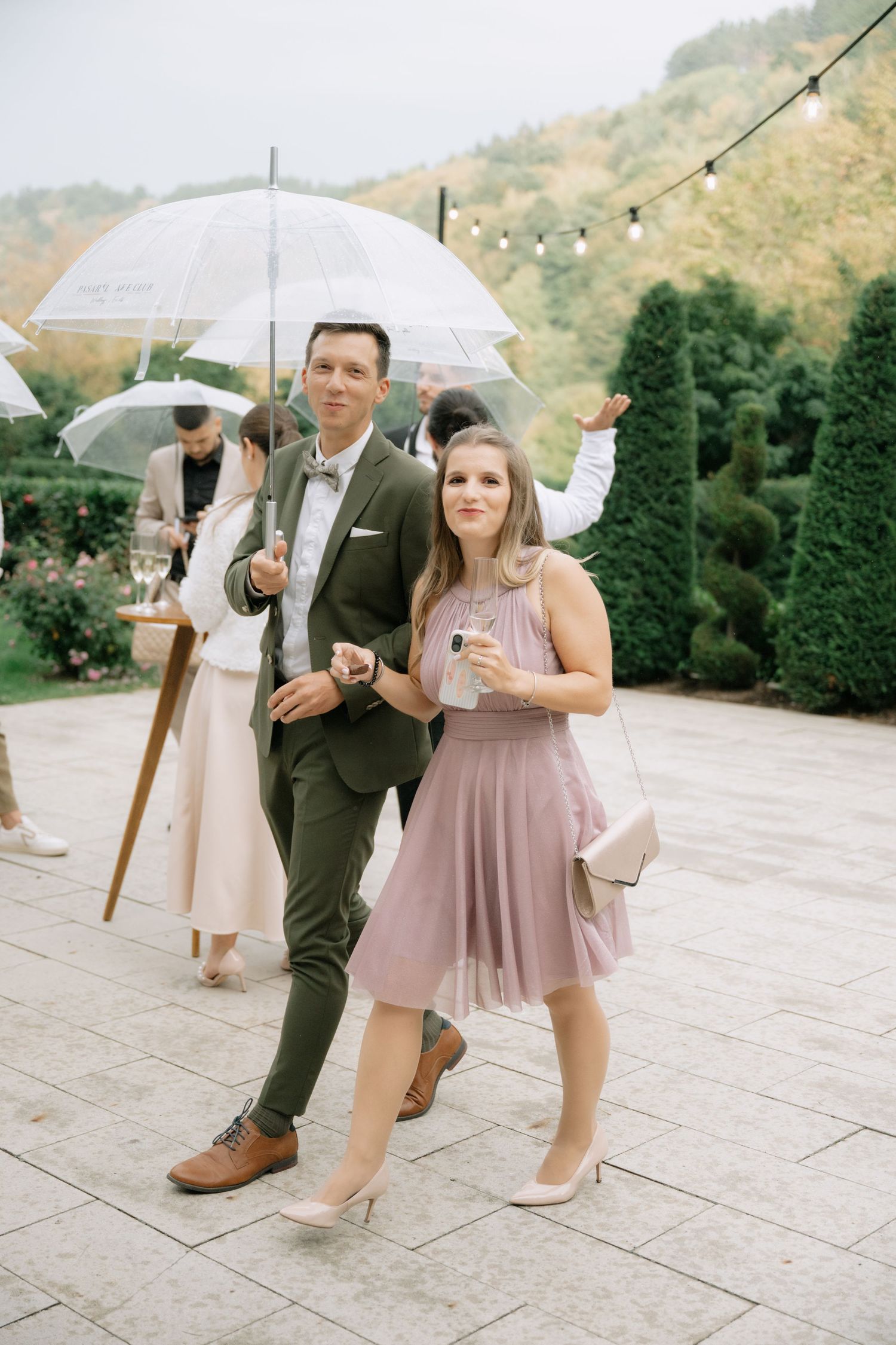 Wedding party members holding white umbrellas walk in a line on stone patio during outdoor celebration in mountains.
