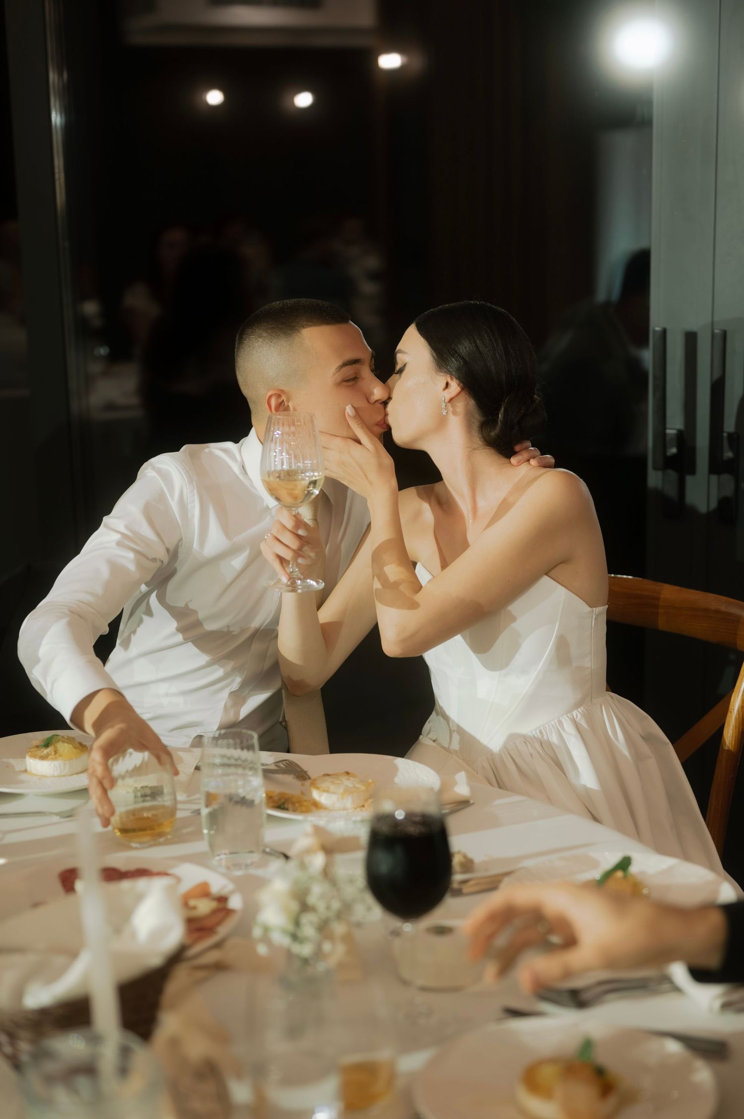 A romantic couple in white attire shares intimate moments over dinner at a dimly lit restaurant table.