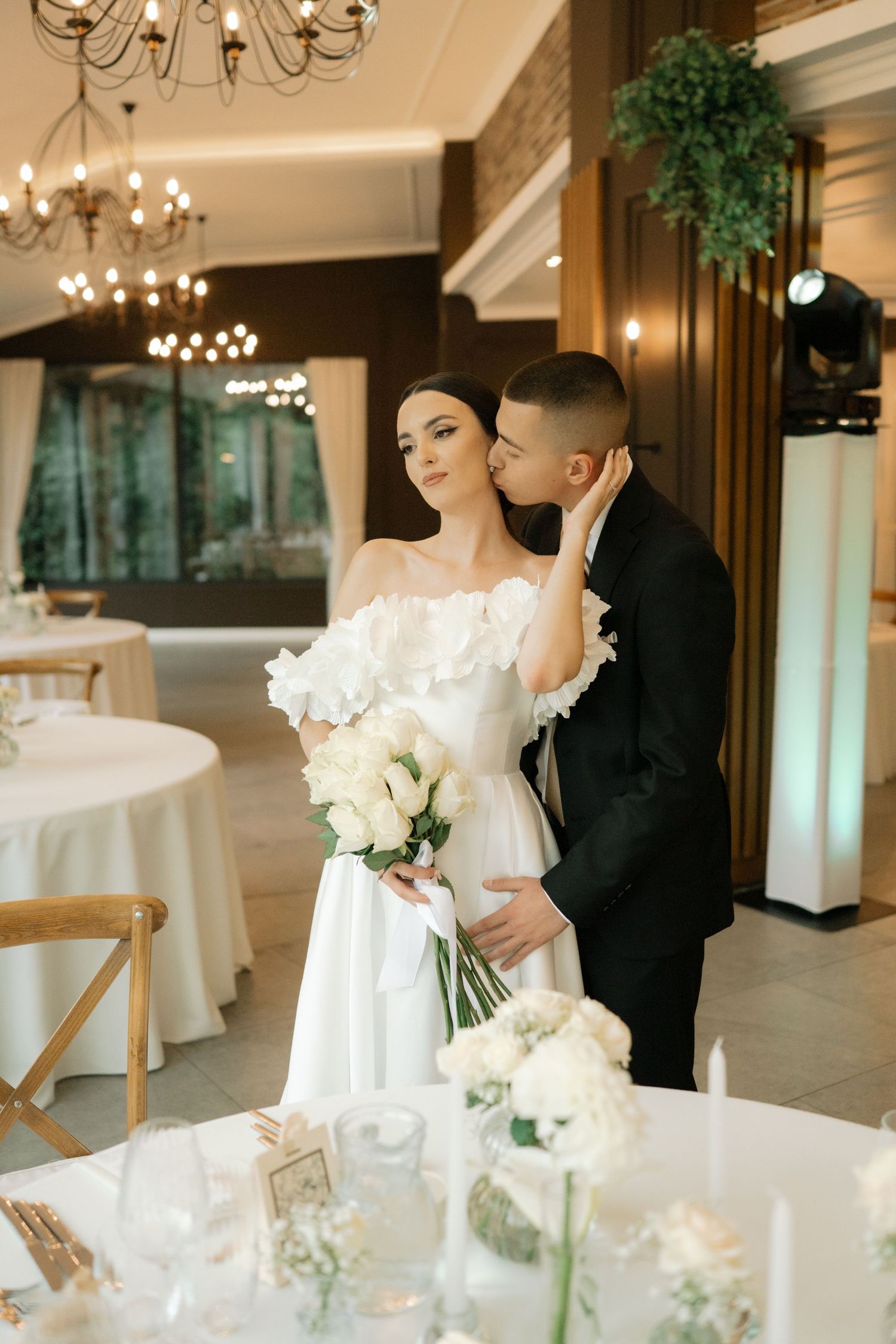 Elegant wedding reception venue with chandeliers and white floral centerpieces as newlyweds share intimate moments.