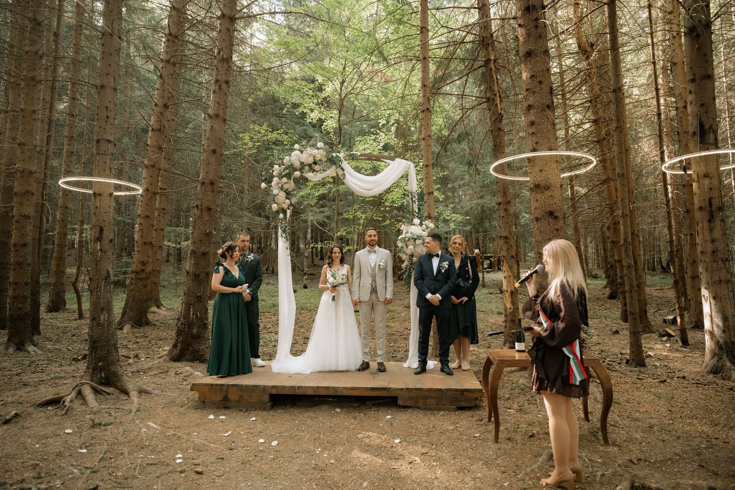 Outdoor forest wedding ceremony on wooden platform with couple, officiant, and wedding party among tall pine trees.