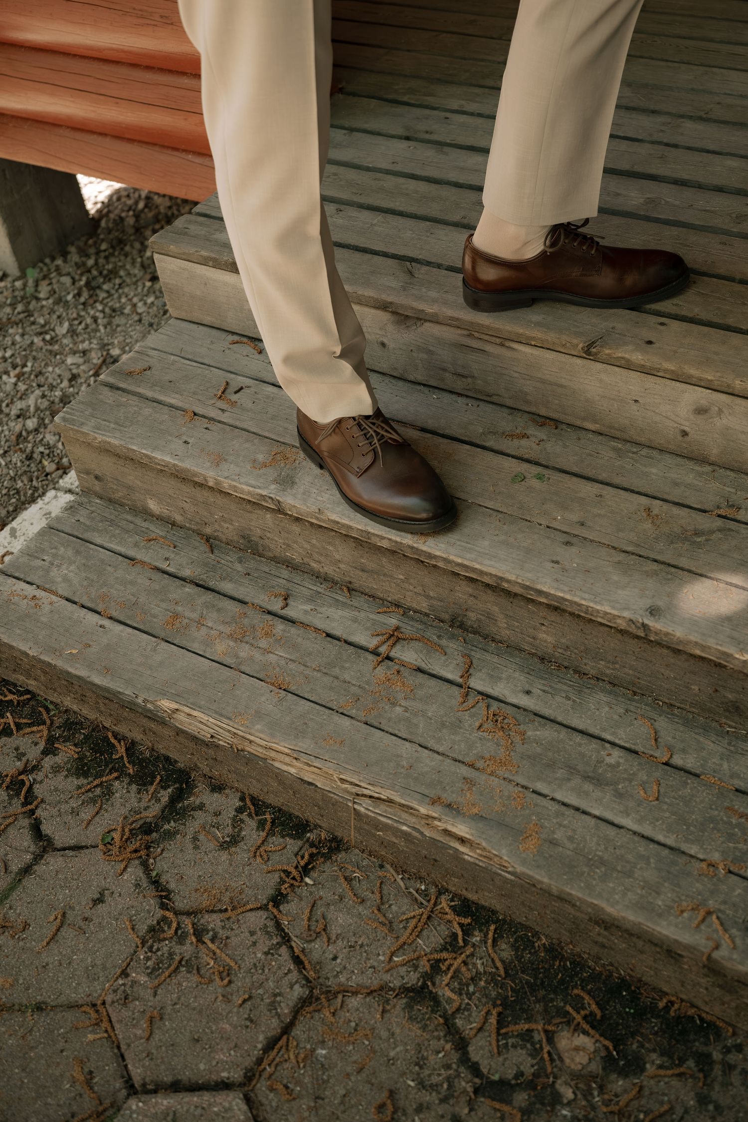 Man wearing beige trousers and brown leather Oxford shoes standing on rustic wooden steps outdoors.