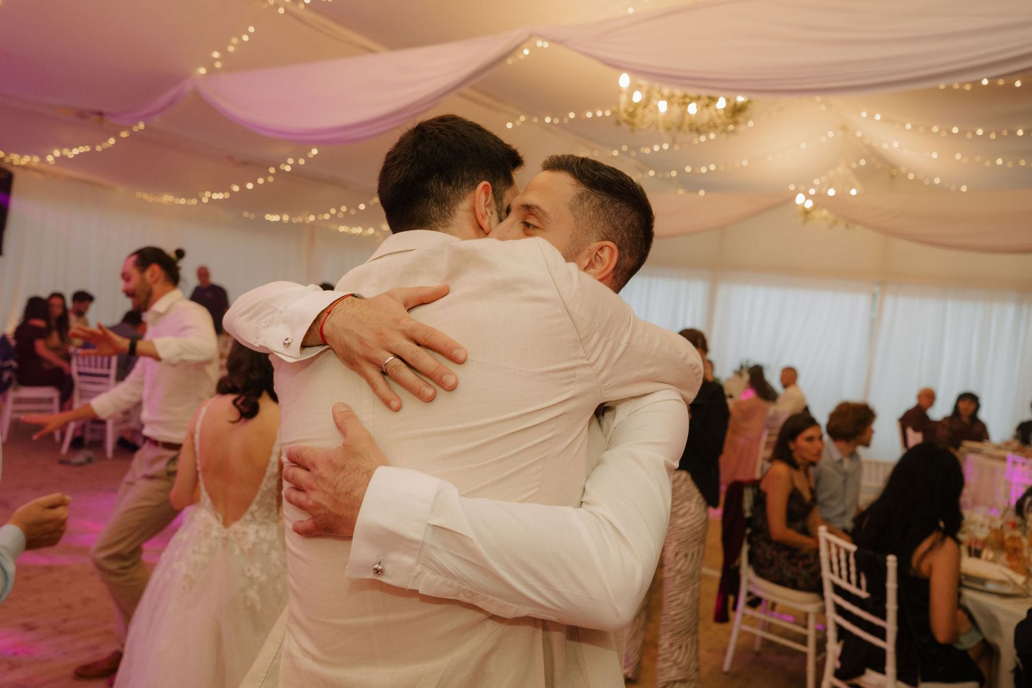 Two grooms share an emotional embrace on the dance floor during their wedding reception under fairy lights.