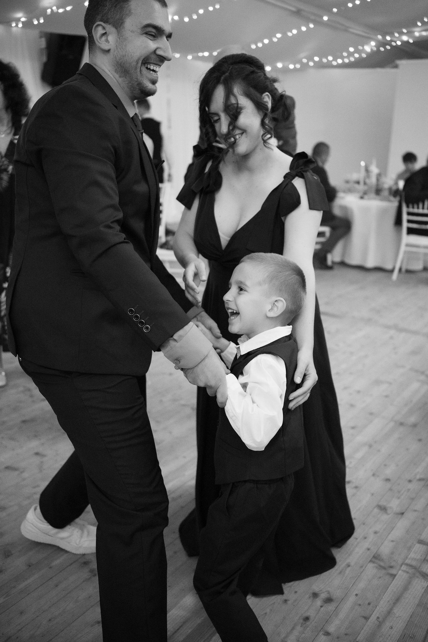 Black and white photo of a couple dancing joyfully with a young boy in formal attire at a wedding reception.