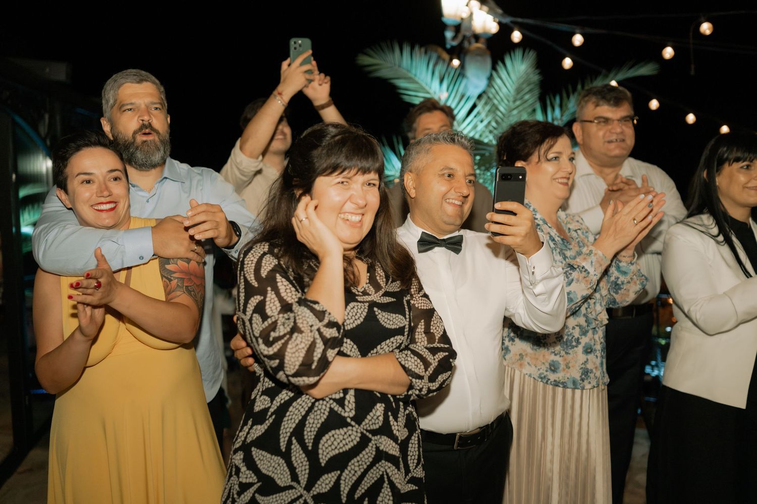 Party guests celebrate with drinks and smiles under string lights at an outdoor evening event.