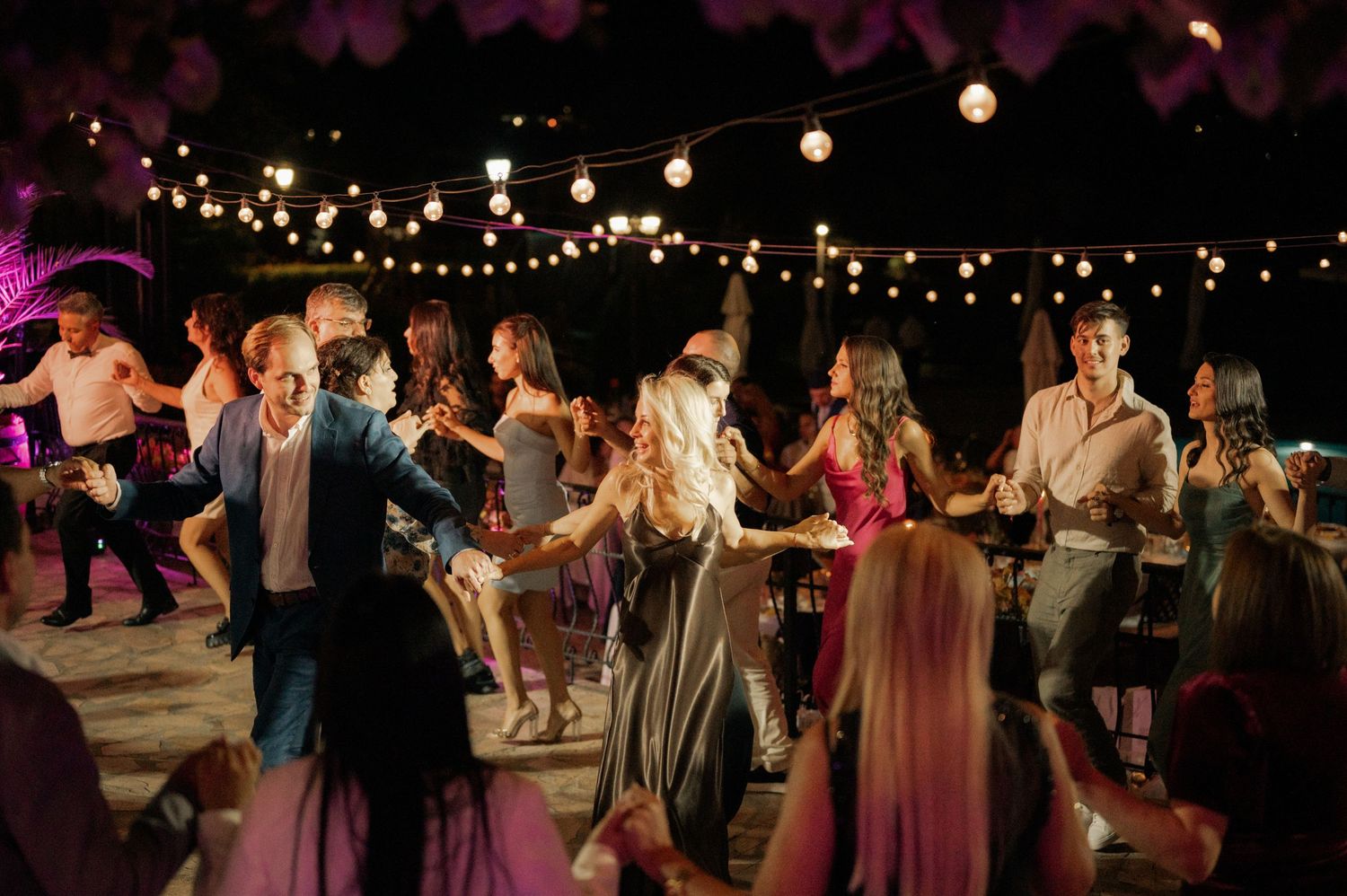 Wedding guests dance under twinkling string lights at an outdoor evening reception celebration.