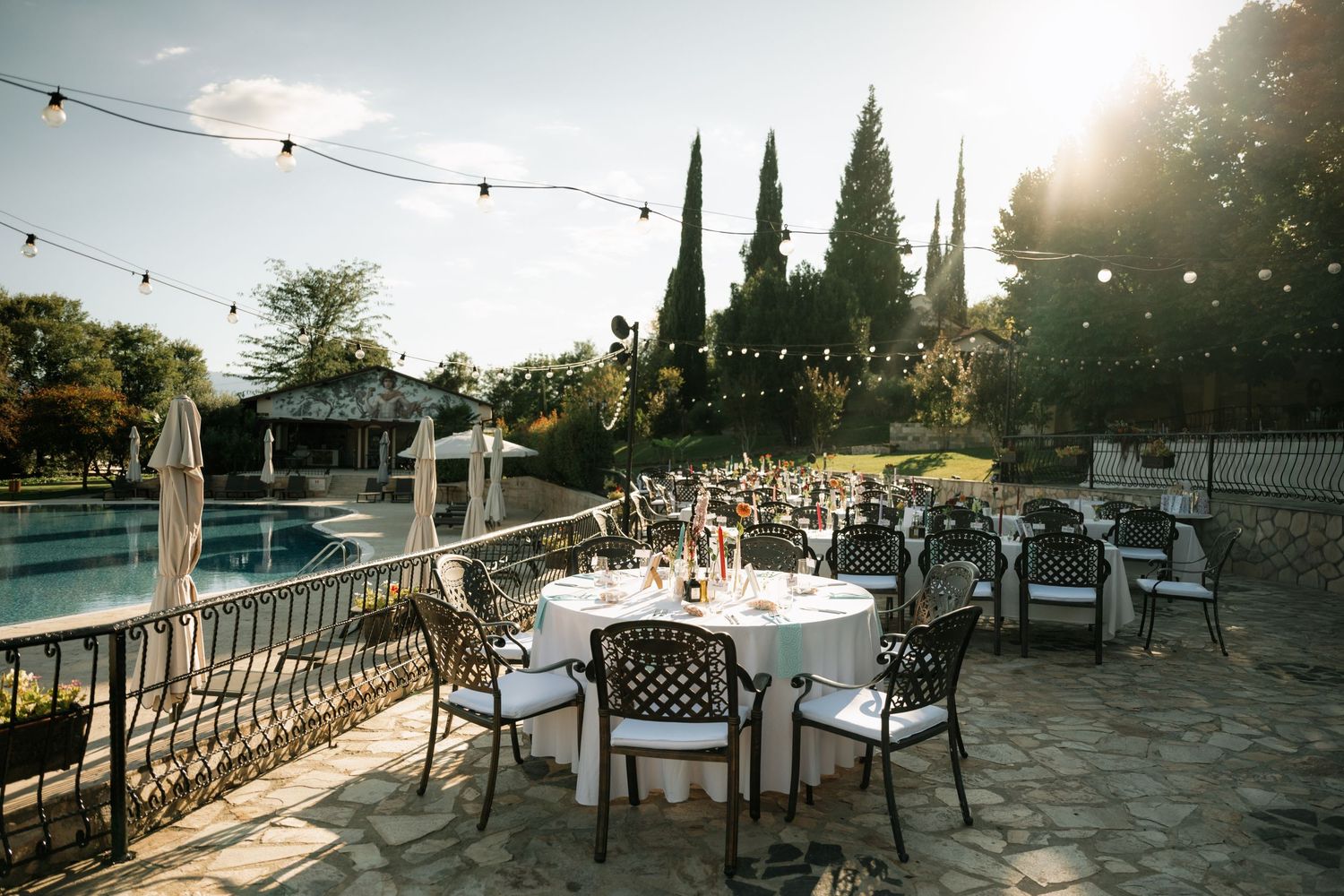 String lights illuminate an outdoor wedding reception venue at sunset with rustic wooden tables and chairs.