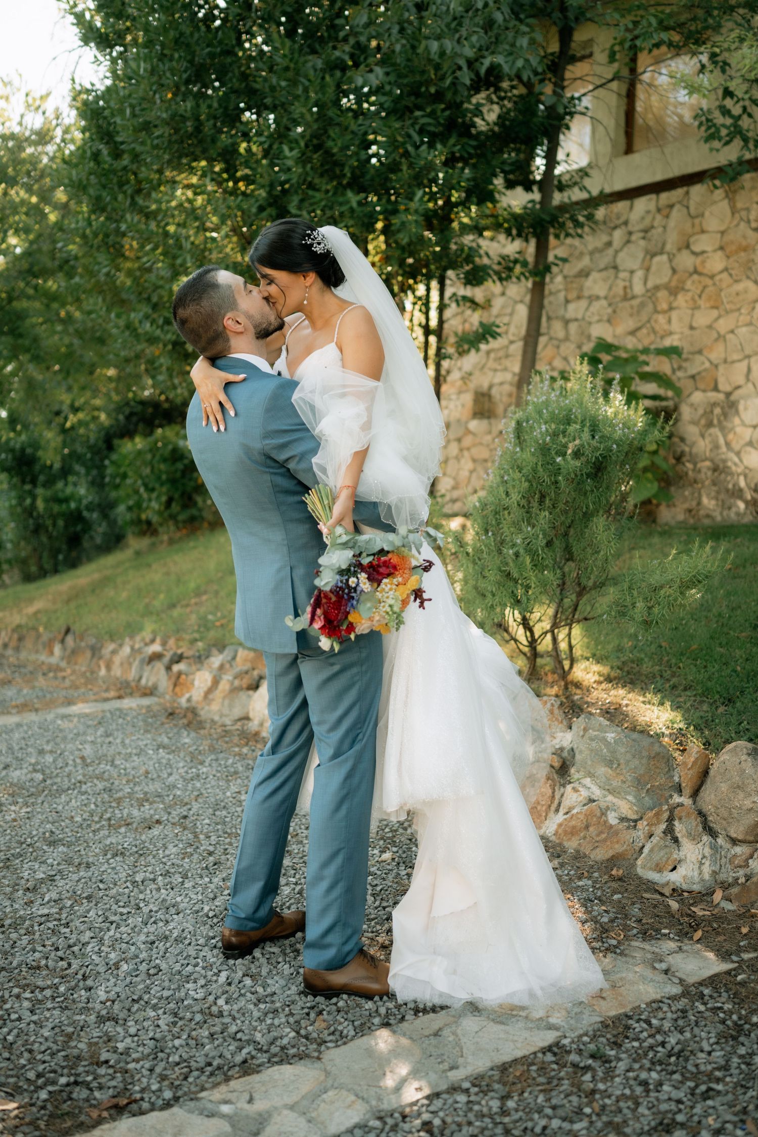 Romantic wedding couple shares an intimate moment on a gravel path against a rustic stone wall at sunset.