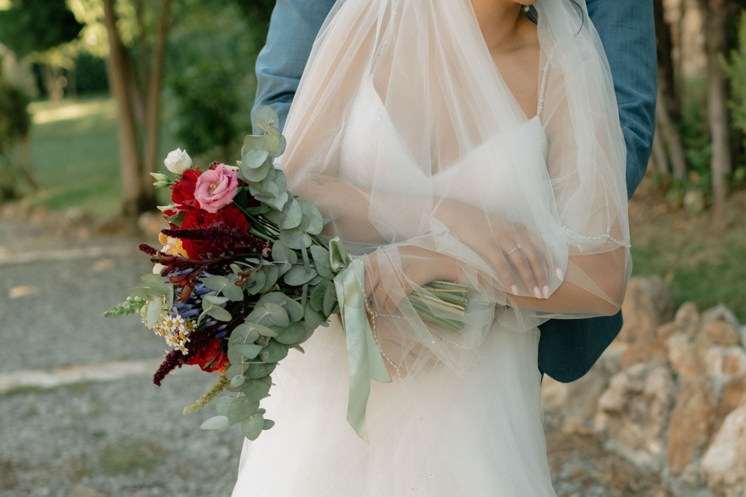 A couple in formal wedding attire poses in a romantic garden setting with soft natural lighting and floral bouquet.
