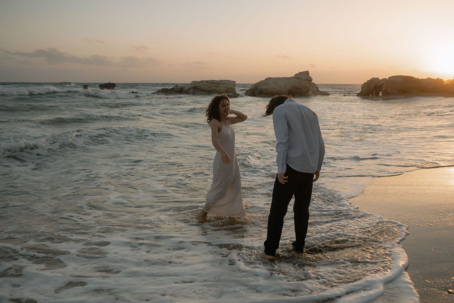 Couple standing in ocean waves at sunset on a rocky beach, woman in white dress and man in white shirt.