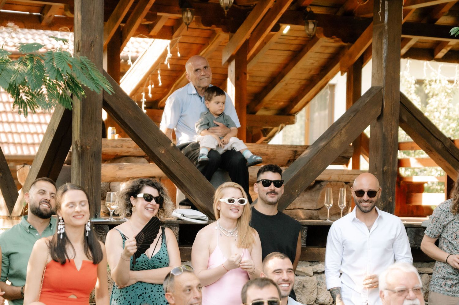 A group of people wearing sunglasses gather under a wooden pavilion structure on a sunny day.