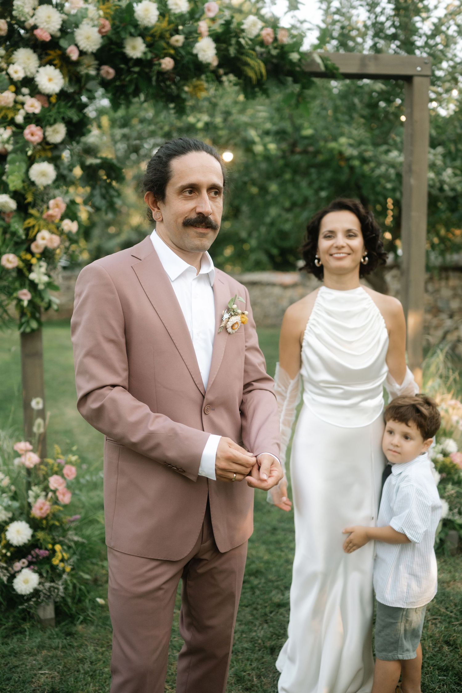 A family poses together in a garden with climbing roses, wearing brown and white formal attire.