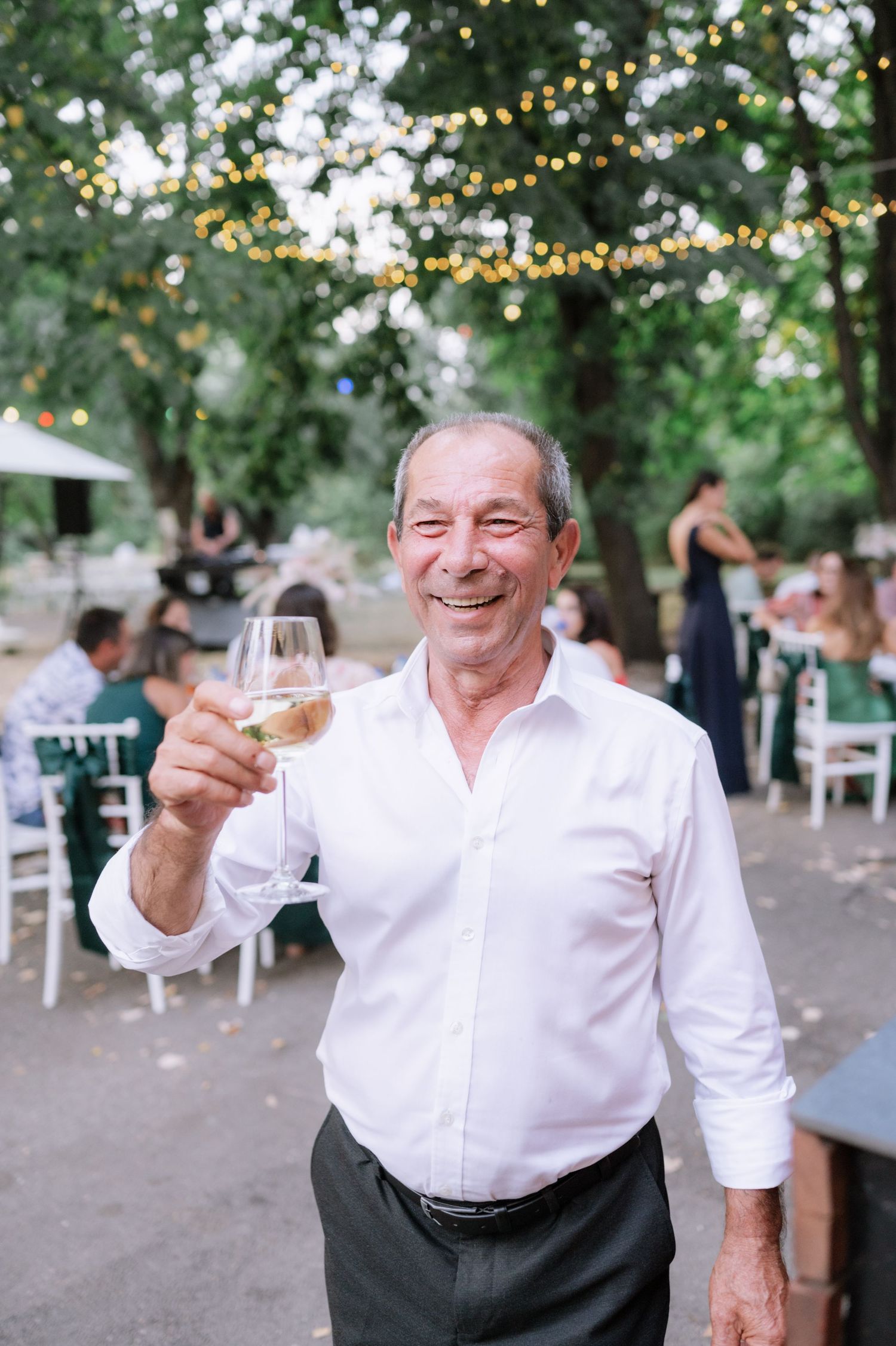 A person in a white shirt enjoys drinks and laughter at an outdoor gathering with string lights overhead.