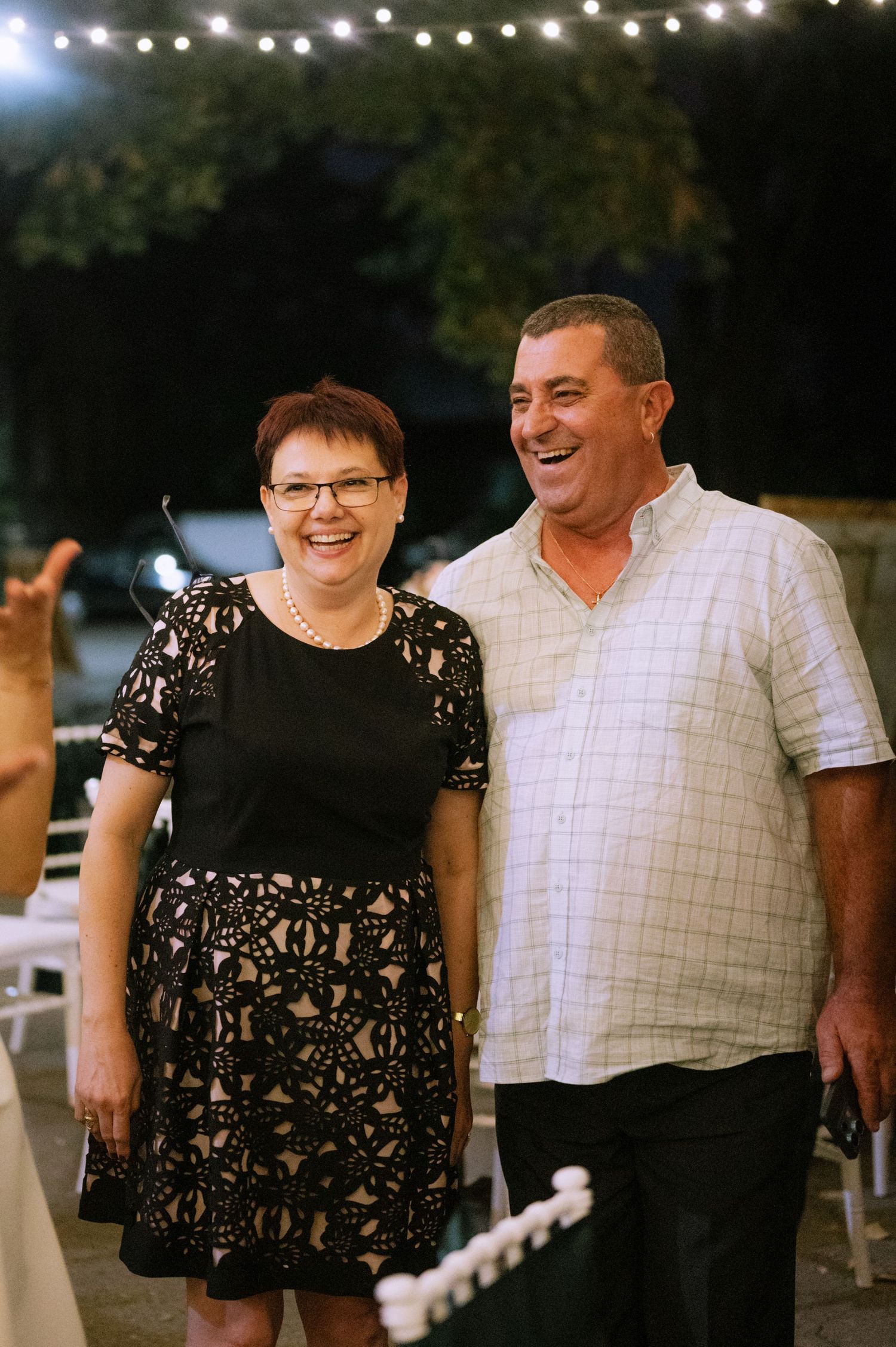 A couple shares joyful laughter together at an outdoor evening event with string lights overhead.