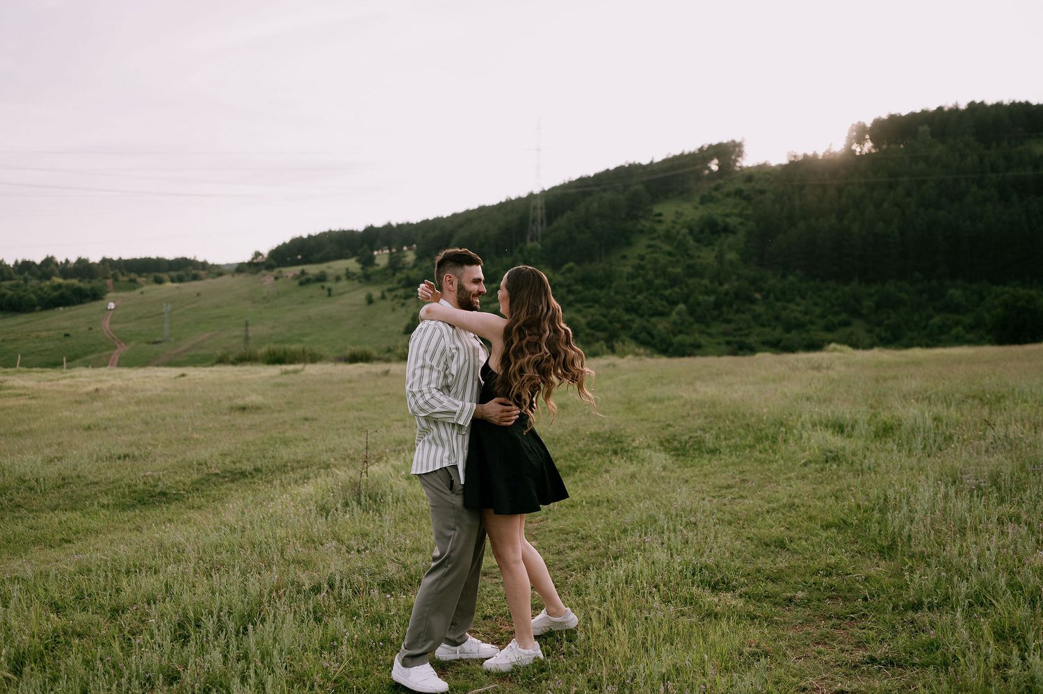 A couple embraces in a romantic moment while standing in a green meadow on an overcast day.