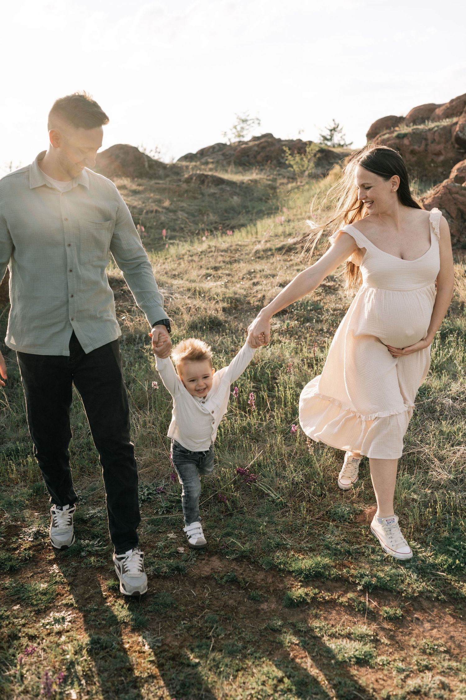 A family holds hands while walking on a grassy hillside at sunset, with the mother wearing a flowing dress.