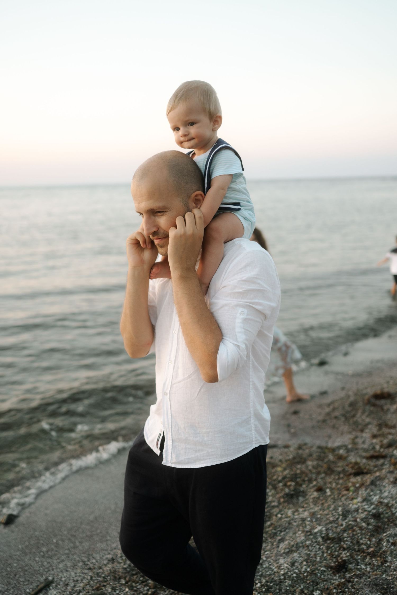 Parent holds baby against sunset sky at rocky beach while waves crash on shore in series of tender moments.