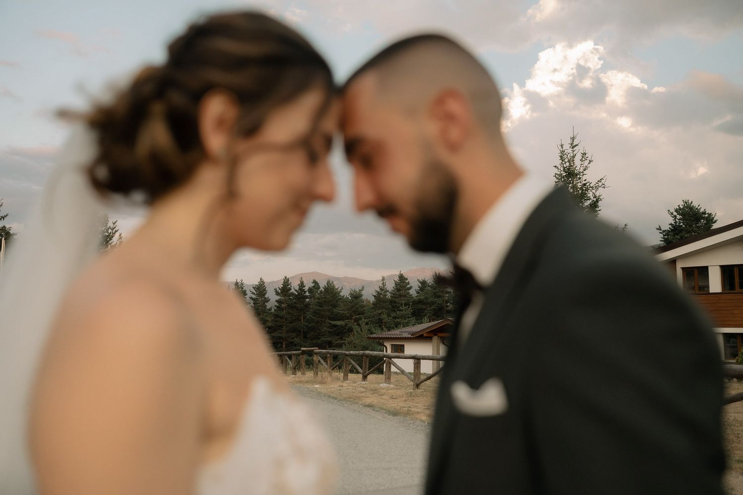 Romantic wedding couple shares an intimate moment at sunset with evergreen trees and a rustic barn in the background.