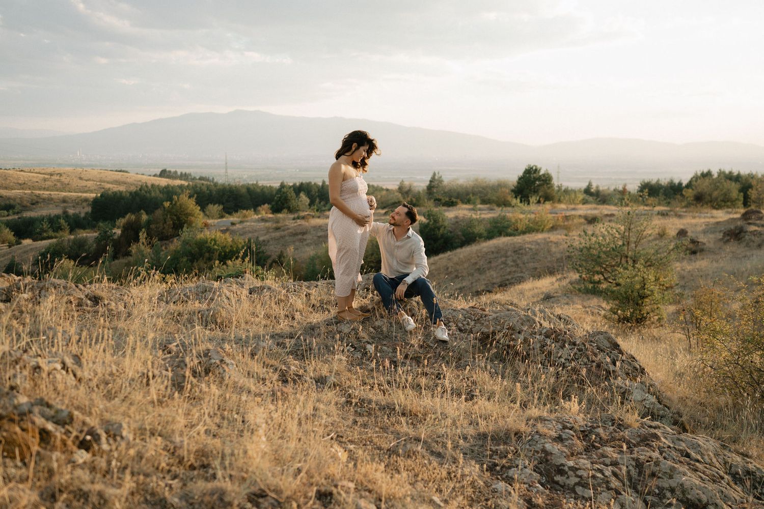 Couple walking together on a golden hillside at sunset with mountains in the distance.