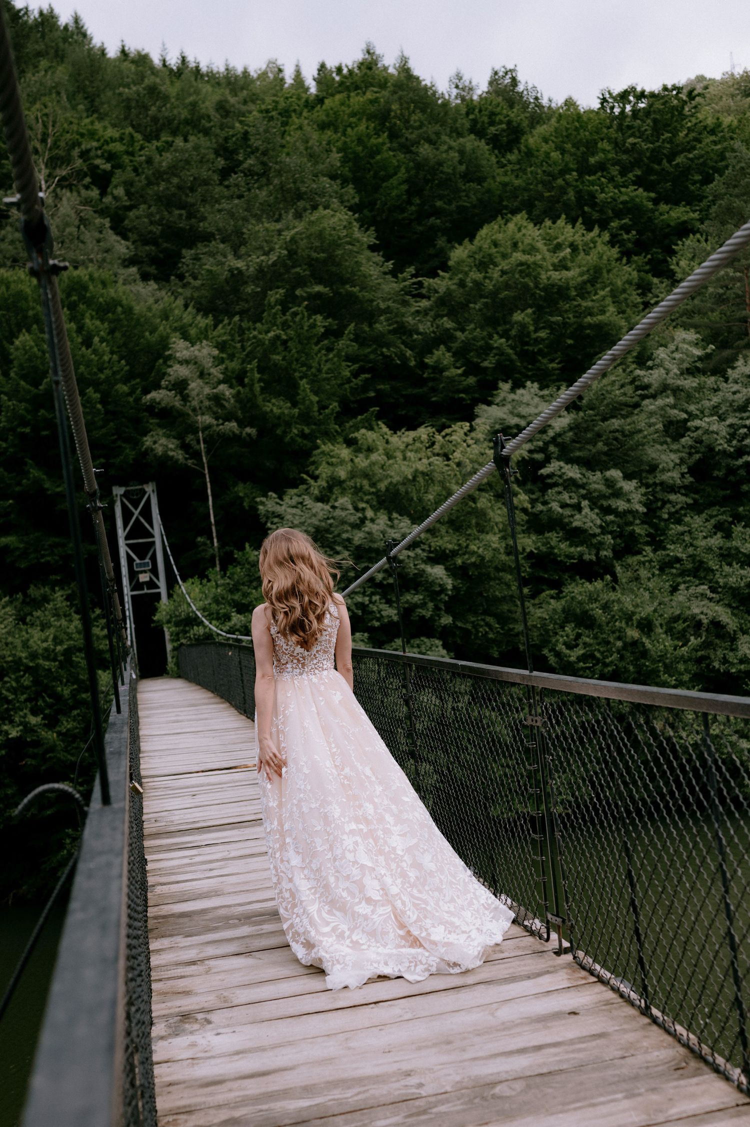 A person in a flowing white lace dress walks across a scenic suspension bridge surrounded by lush forest canopy.
