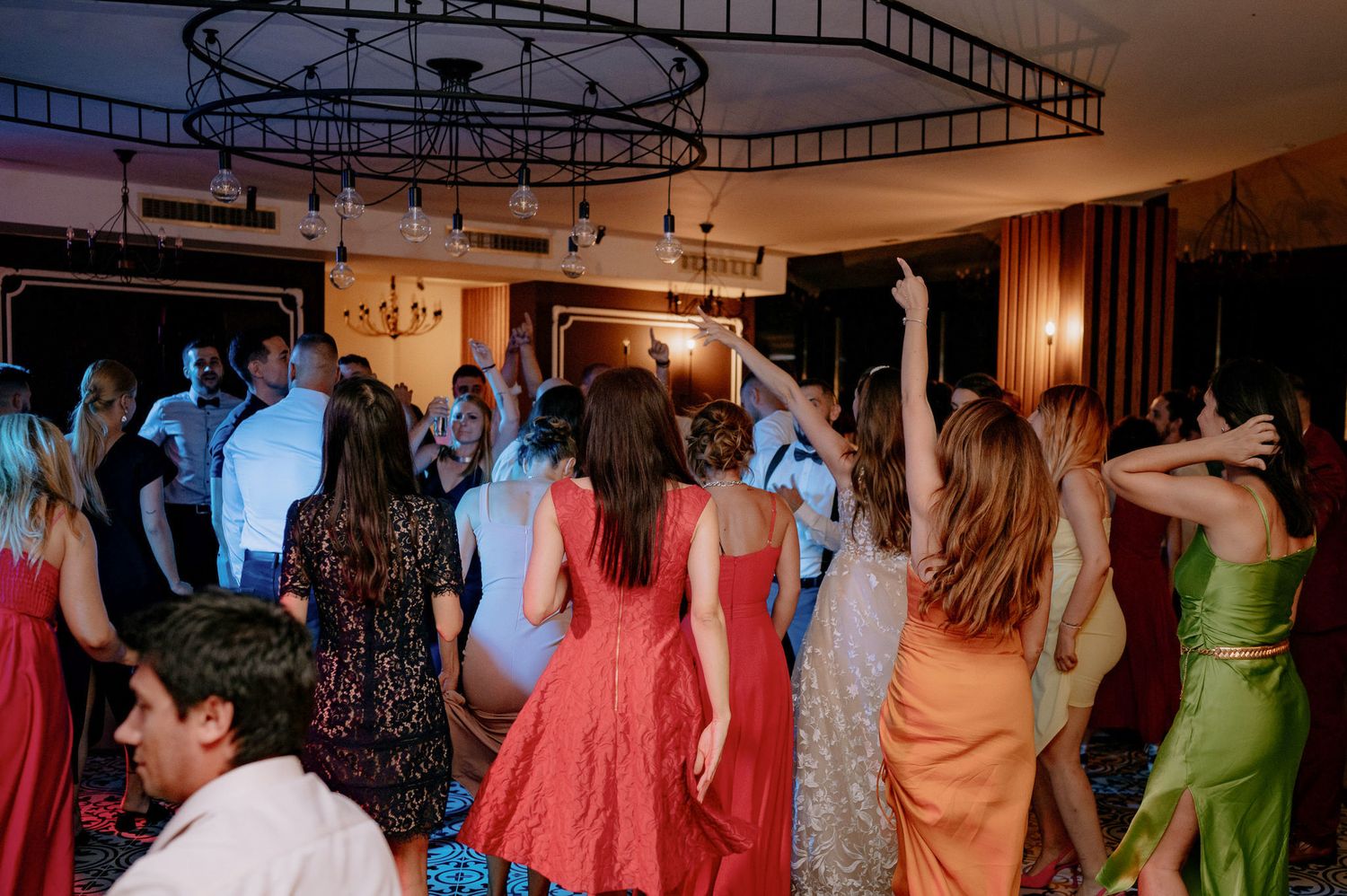 Wedding guests dance and celebrate on a dimly lit dance floor surrounded by colorful party lights and decorations.