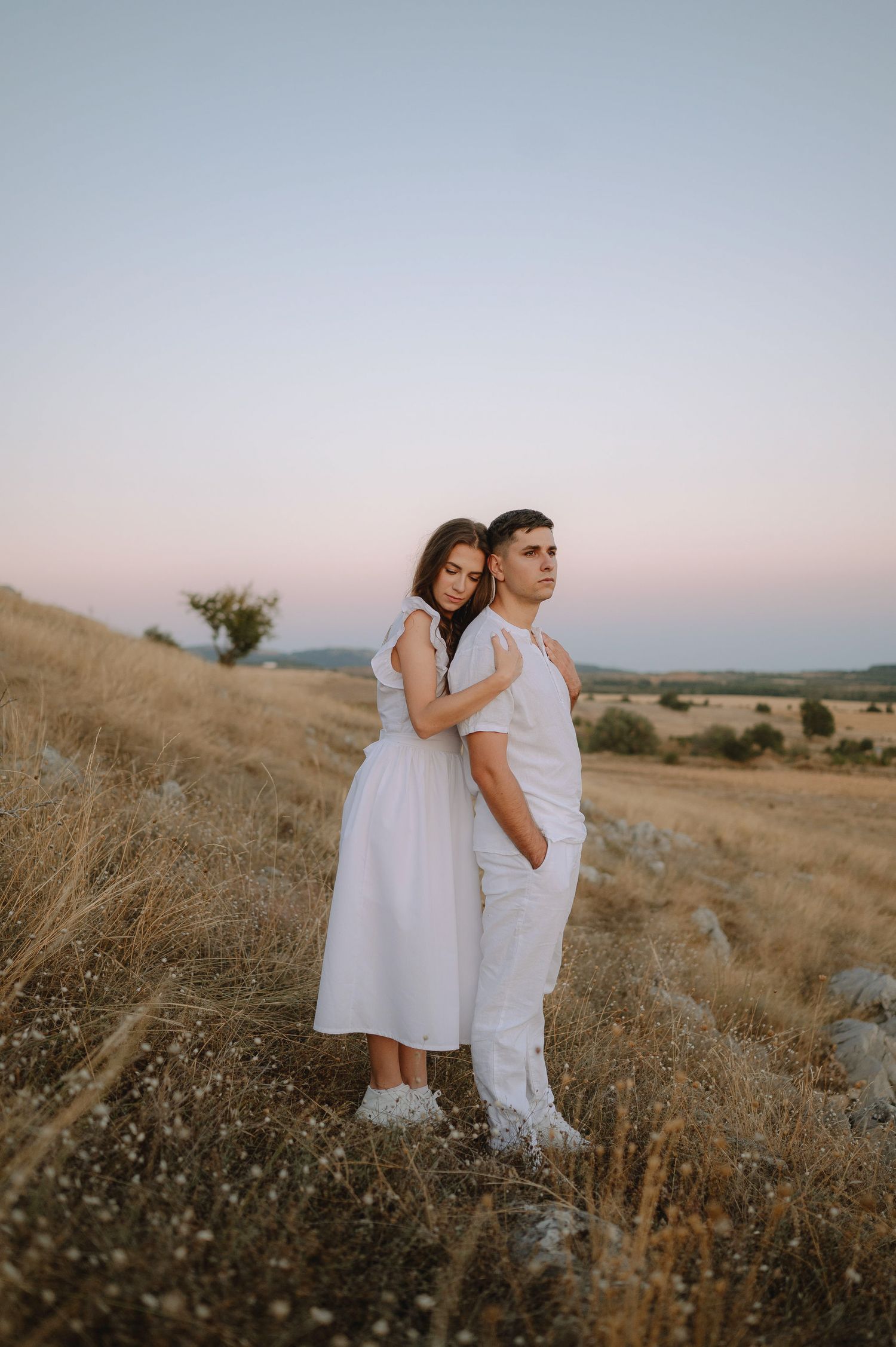 A couple in white outfits stands together in a golden field at sunset creating a romantic silhouette against the sky.