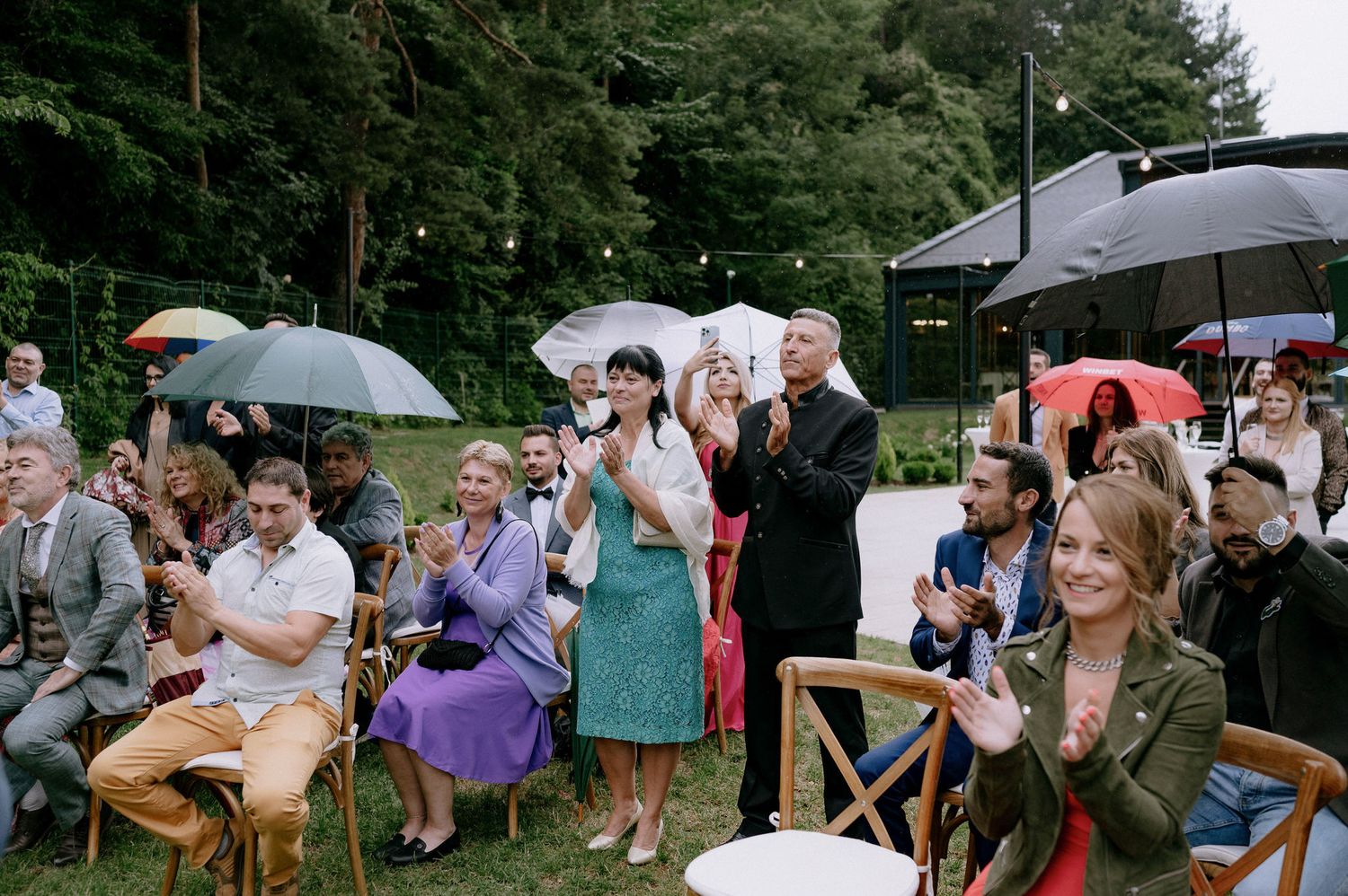 A rainy outdoor wedding ceremony with guests holding umbrellas while seated in white chairs among trees.