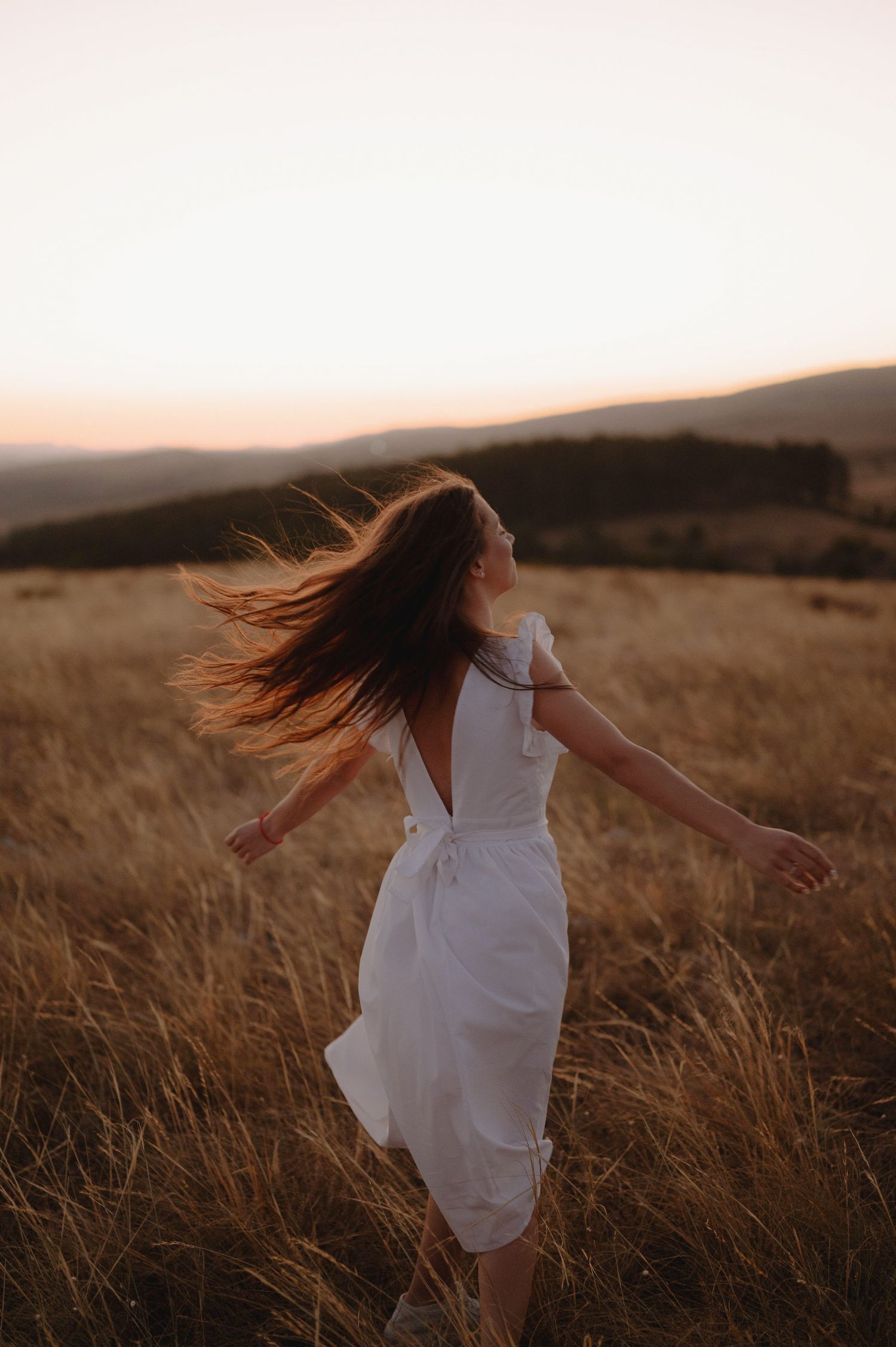 A figure in a flowing white dress dances through a golden wheat field at sunset in the countryside.